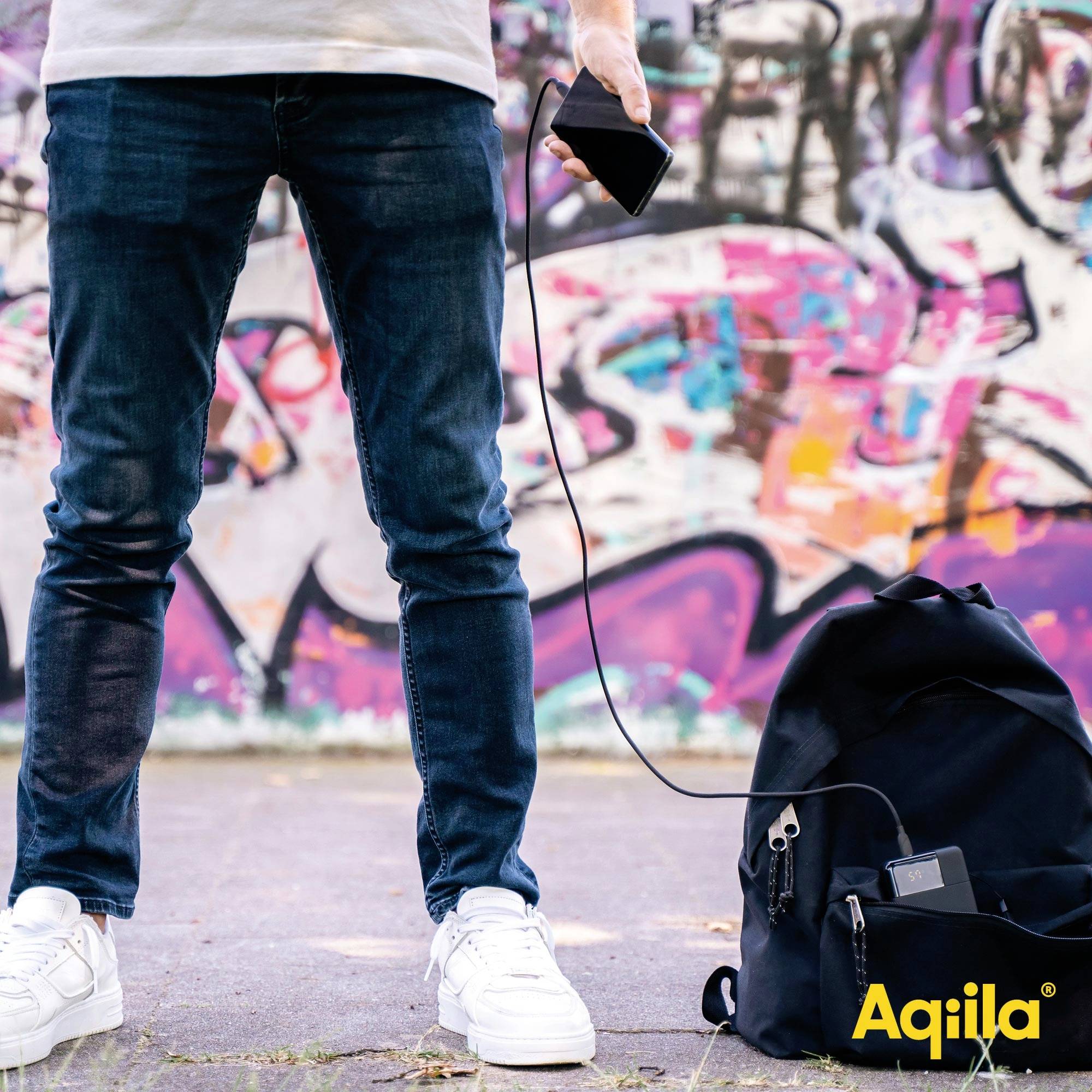 Osoba trzyma telefon komórkowy, stojąc przed kolorową ścianą pokrytą graffiti. Na ziemi leży plecak, podłączony do kabla ładującego.