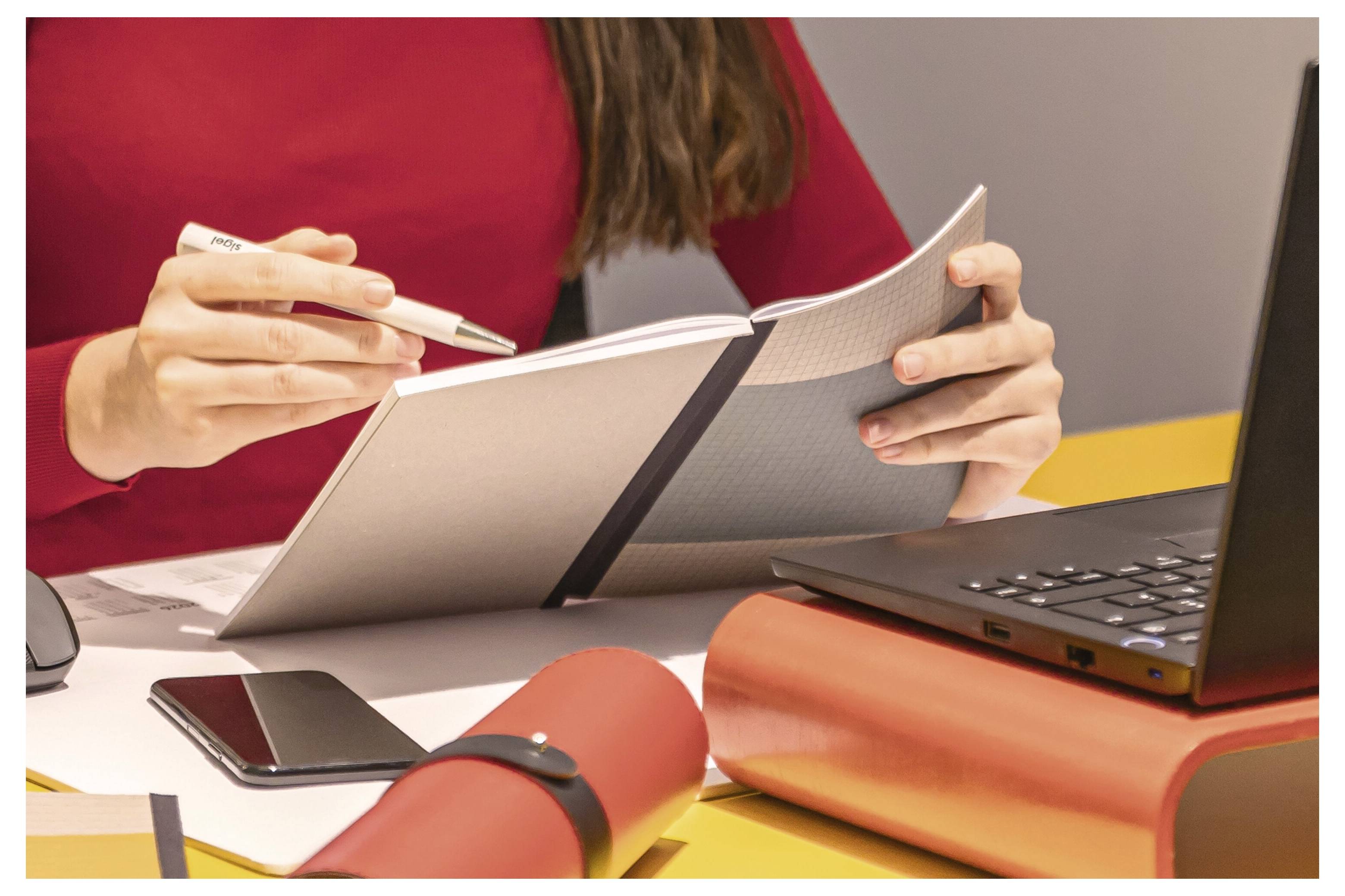 Osoba w czerwonym topie pisze w notesie obok laptopa, ze smartfonem i zrolowanymi papierami na biurku.