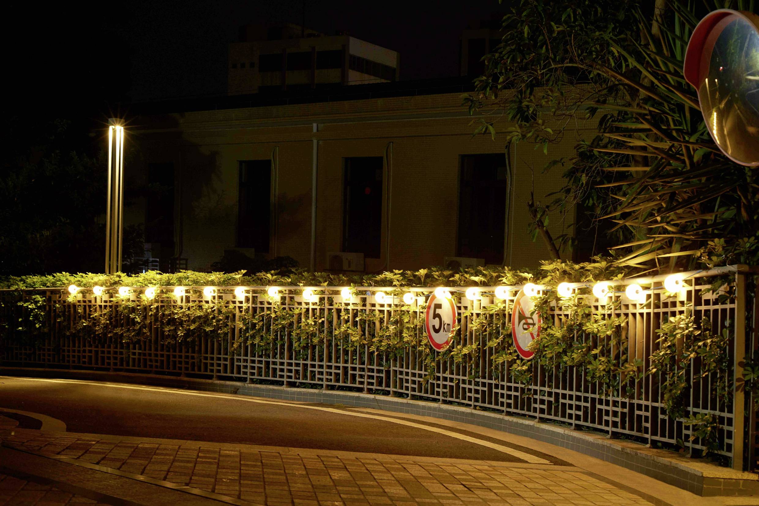 Oświetlona ulica nocą z łańcuchem świetlnym na ogrodzeniu, znakami drogowymi '5' i '2' km/h oraz oświetlonym chodnikiem.