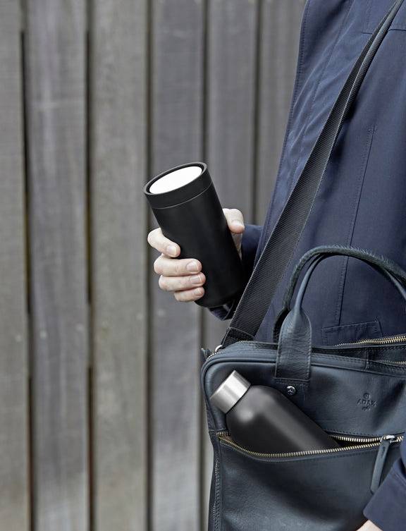 Osoba trzyma czarny kubek i ma torbę z butelką przewieszoną przez ramię. W tle: drewniana ściana.