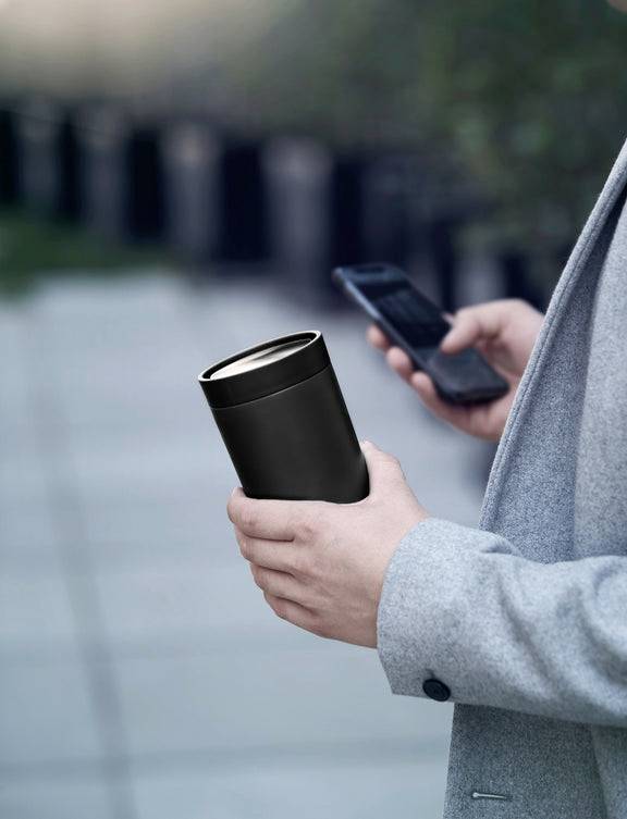 Osoba trzyma czarny termokubek w lewej ręce i smartfon w prawej ręce, spacerując na dworze.