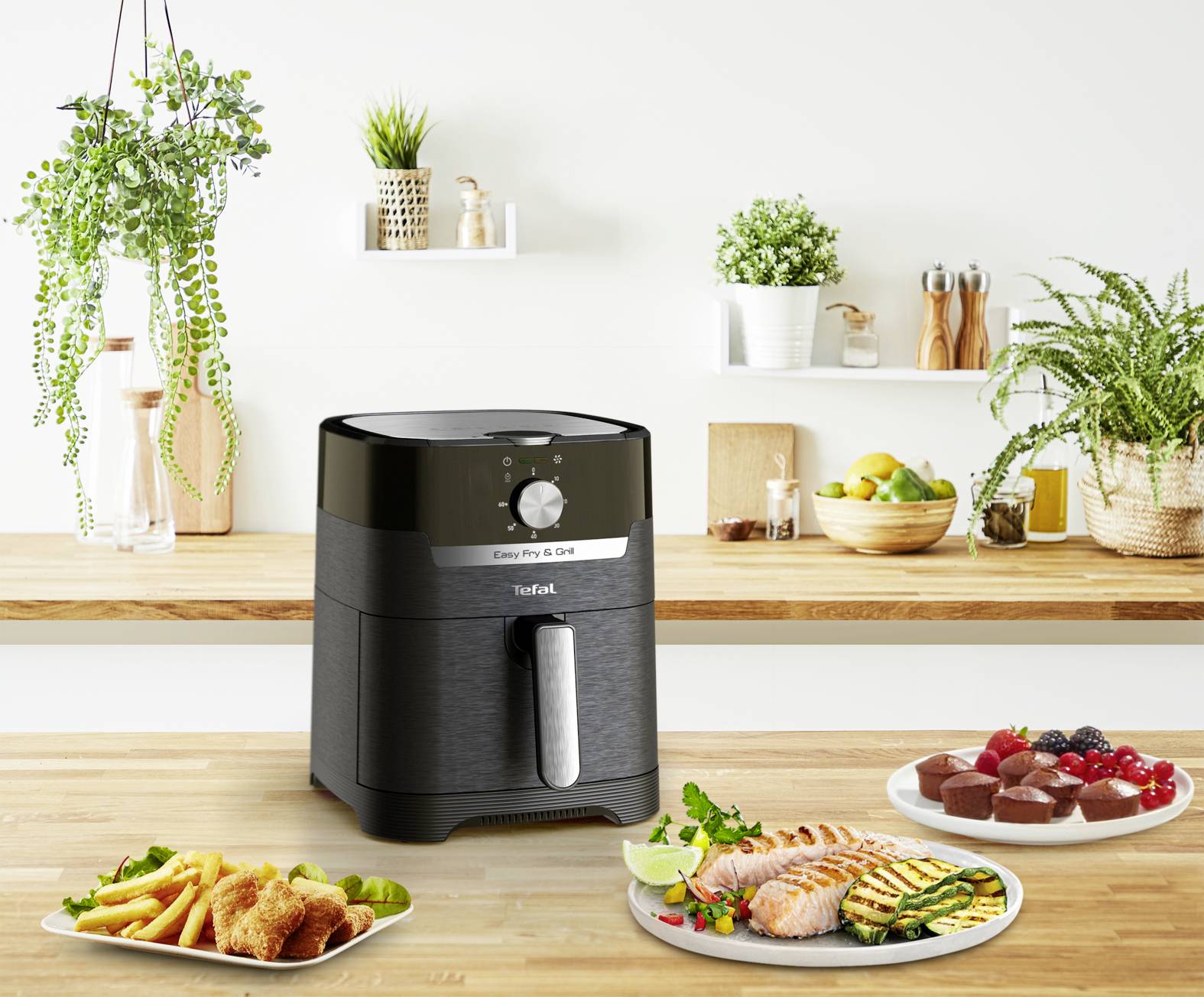 Na blacie kuchennym stoi czarna frytkownica beztłuszczowa, otoczona roślinami i przyborami kuchennymi. Na pierwszym planie znajdują się talerze z potrawami smażonymi, łososiem i deserami.