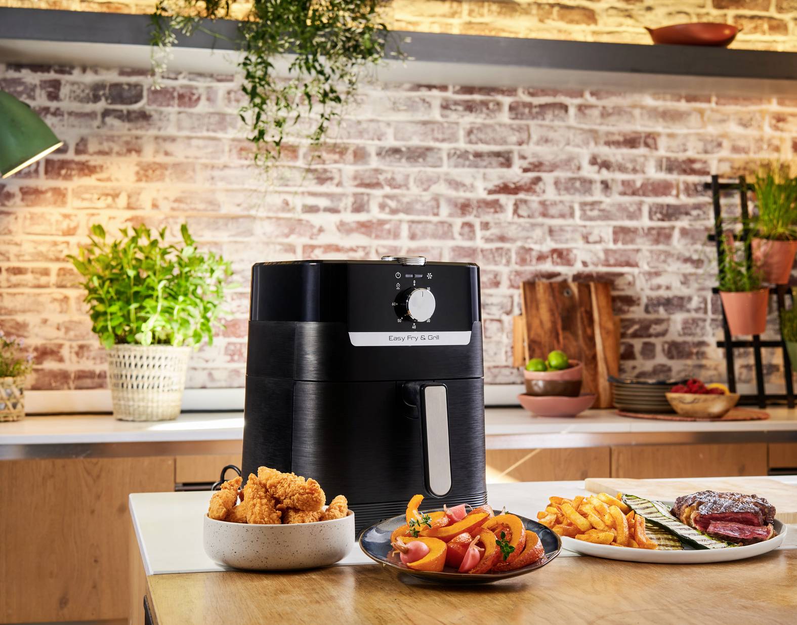 Czarna frytownica na gorące powietrze umieszczona na blacie kuchennym, otoczona talerzami z frytkami, kurczakiem i warzywami. W tle: ściana z czerwonej cegły.