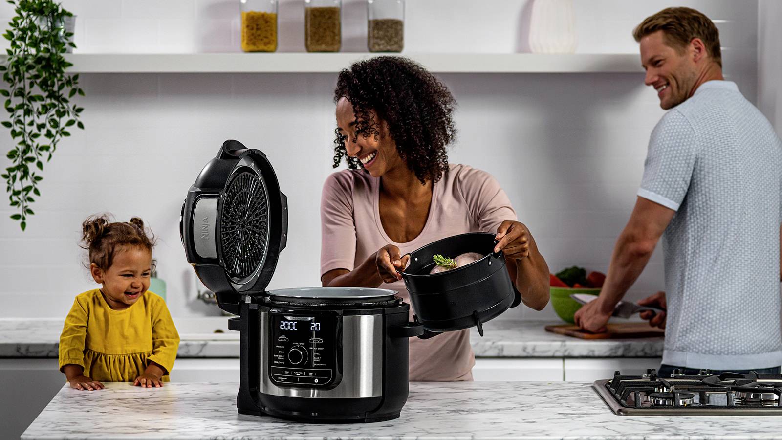 Kobieta gotuje w garnku szybkowarowym w kuchni, podczas gdy małe dziecko przypatruje się, a mężczyzna w tle kroi warzywa.