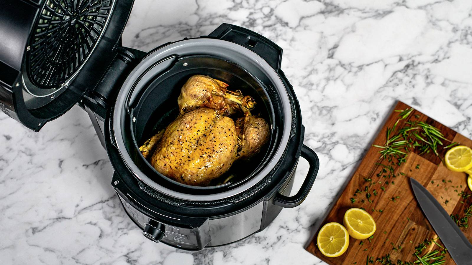 Cały, doprawiony pieczony kurczak leży w szybkowarze na marmurowym blacie, obok leży deska do krojenia z ziołami i cytrynami.