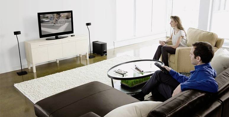 Mężczyzna i kobieta siedzą w salonie na sofach i oglądają telewizję. Pomieszczenie jest urządzone nowocześnie, z jasnym dywanem i meblami.