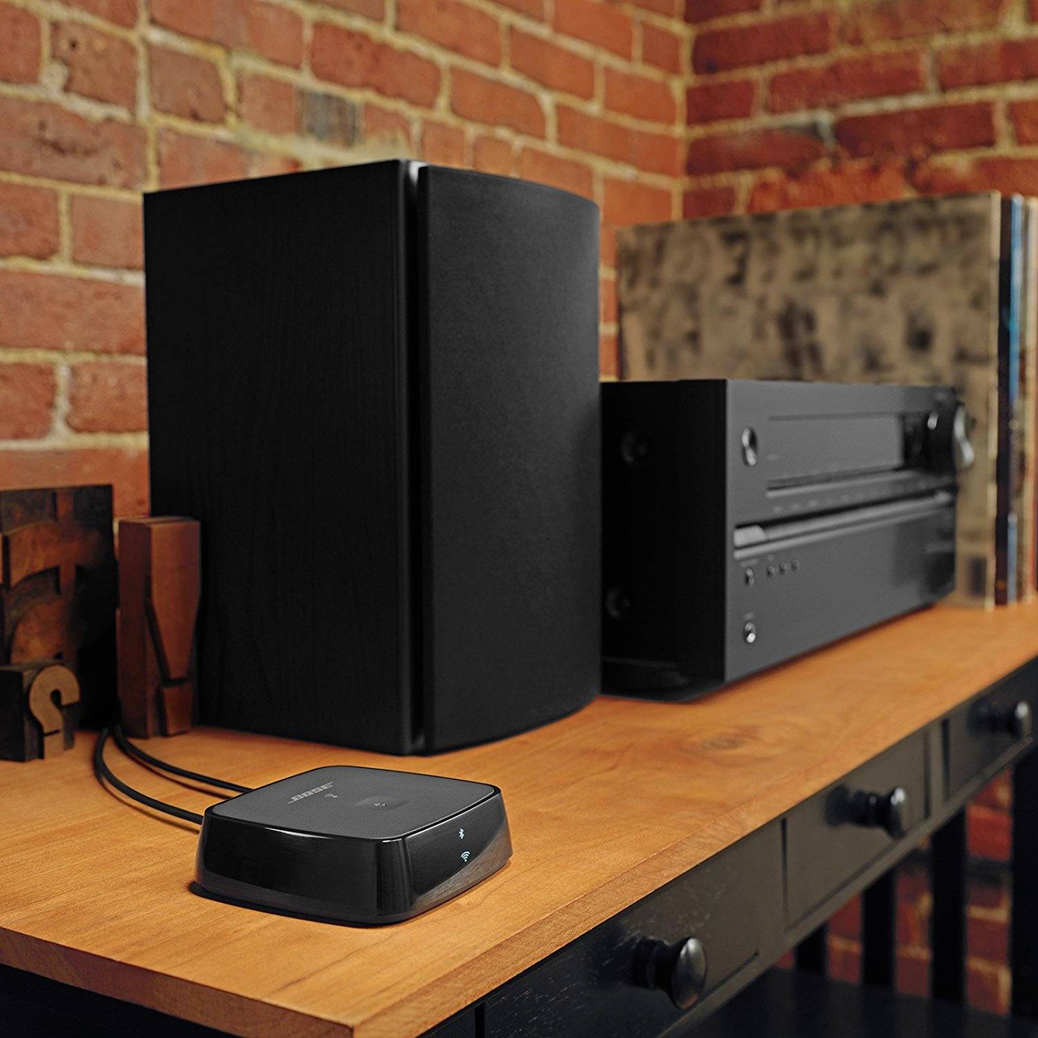 'Głośnik i odbiornik audio stoją na drewnianej półce przed ścianą z czerwonej cegły. Pomieszczenie ma przytulny klimat.'