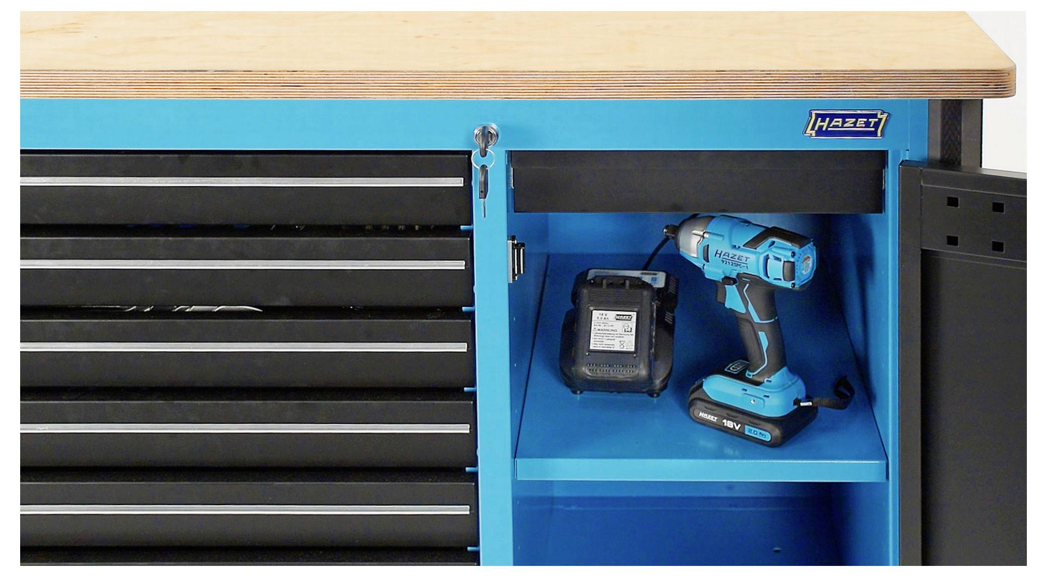 Niebieski szafka narzędziowa z częściowo otwartymi drzwiami i szufladami. Wewnątrz na półce stoi niebieski akumulatorowy wkrętak wraz z ładowarką.