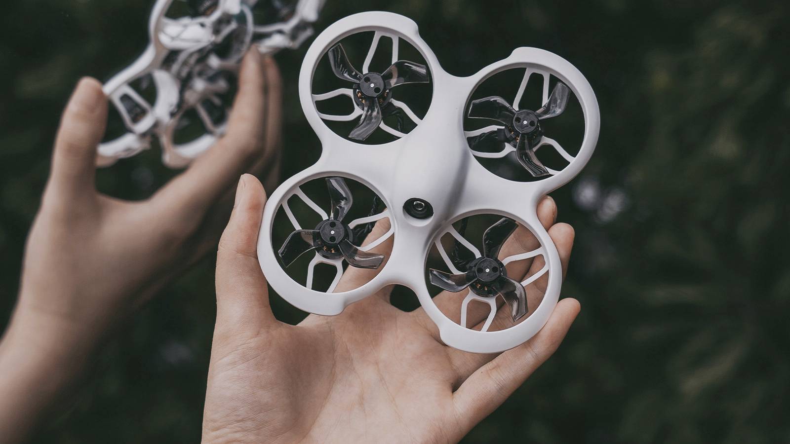 Osoba trzyma małego, białego drona z czterema śmigłami. Dron unosi się tuż nad dłońmi. Tło jest rozmyte.