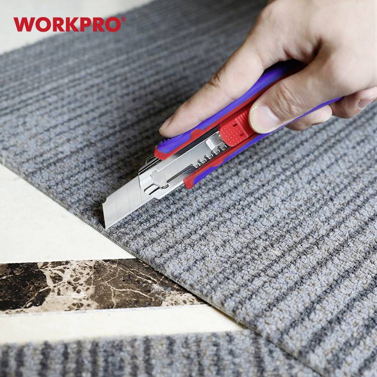 Ręka trzyma nóż do dywanów i przecina szary dywan na jasnym podłożu.