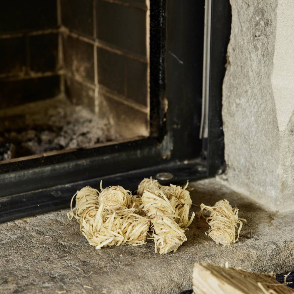 Kilka podpałek z wełny drzewnej leży na kamiennym gzymsie kominkowym obok otwartego kominka z ciemnym tłem.