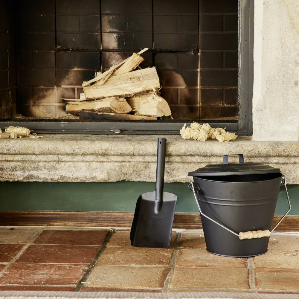 Kominek z ułożonymi kawałkami drewna, gotowymi do rozpalenia. Obok stoi czarny kubeł z łopatką, na podłodze wyłożonej cegłami.