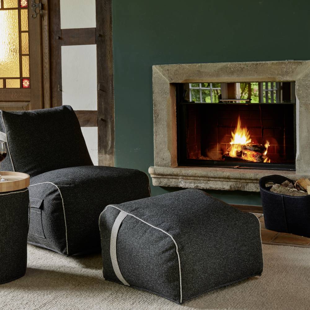 Dwa szare fotele i stołek stoją przed palącym się kominkiem w przytulnym pokoju z elementami drewnianymi i szklaną bramą.