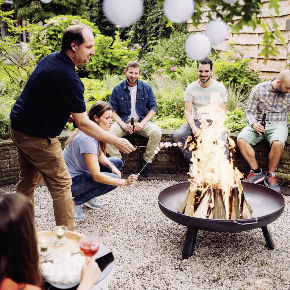 Grupa ludzi siedzi wokół ogniska na świeżym powietrzu i piecze pianki marshmallow. W tle widać zieloną roślinność.