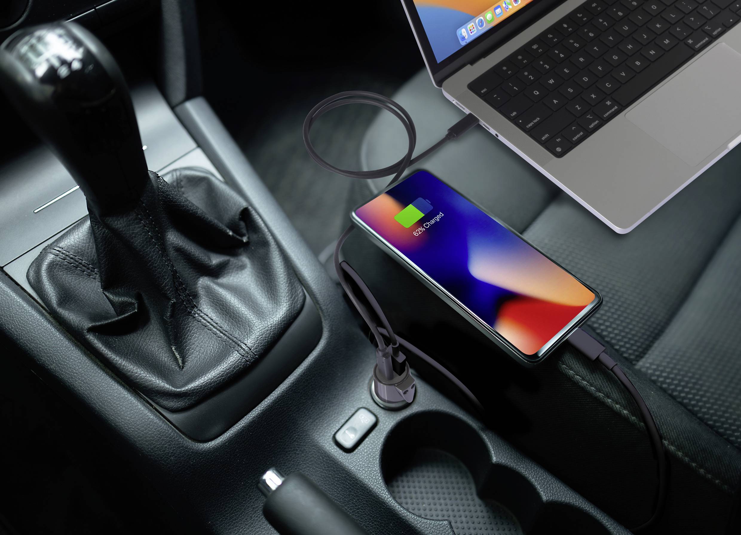 Smartfon ładuje się bezprzewodowo w samochodzie obok skrzyni biegów i laptopa. Ekran wyświetla symbol ładowania.