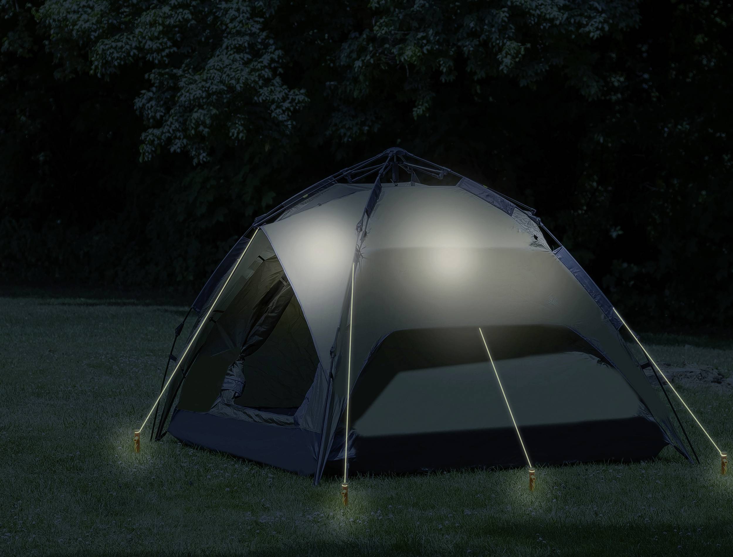 Namiot stoi nocą na łące. Jest oświetlony i otoczony drzewami.
