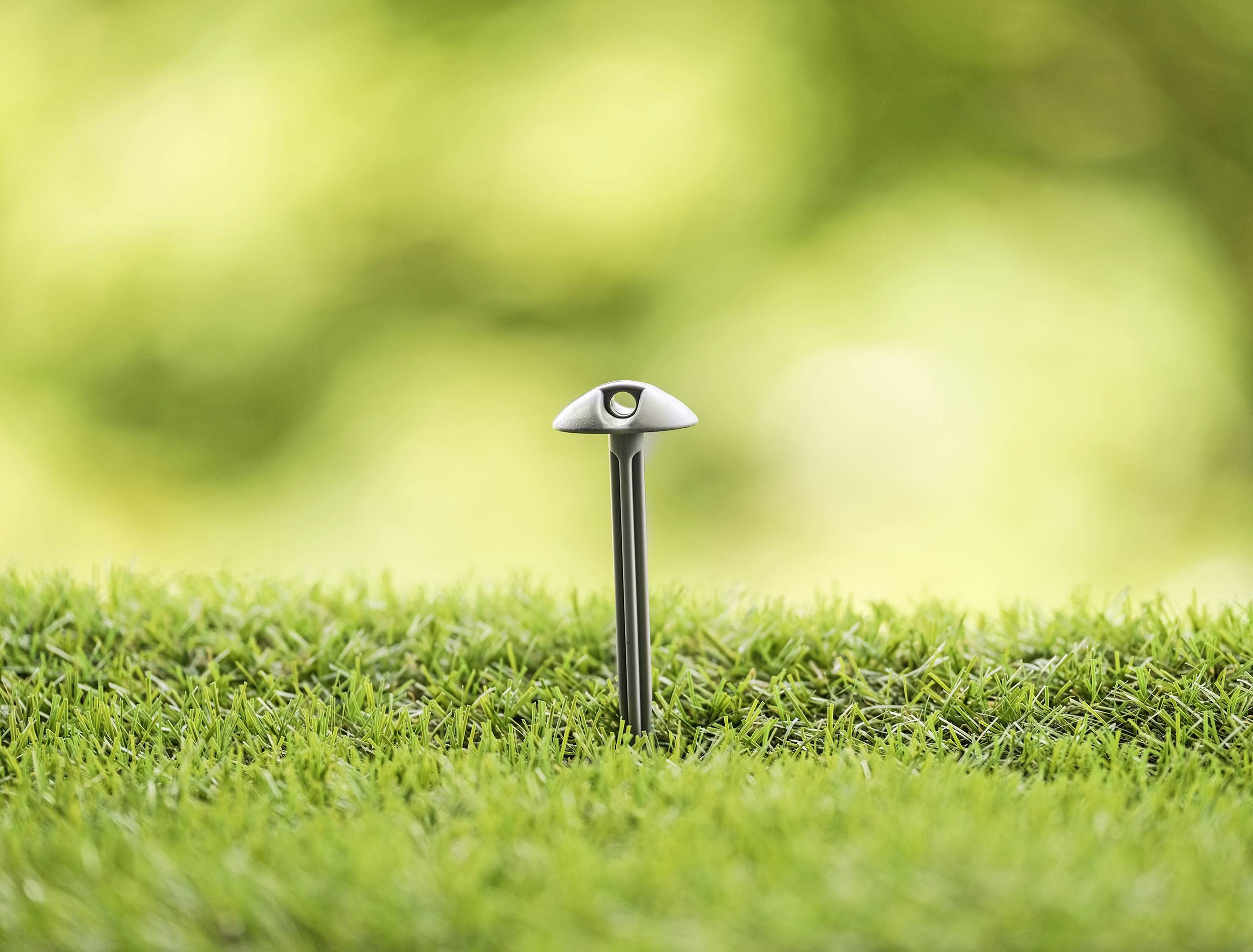 Pojedynczy, nowoczesny słup metalowy z małym elementem świetlnym stoi na gęstym, zielonym trawniku na rozmytym tle.