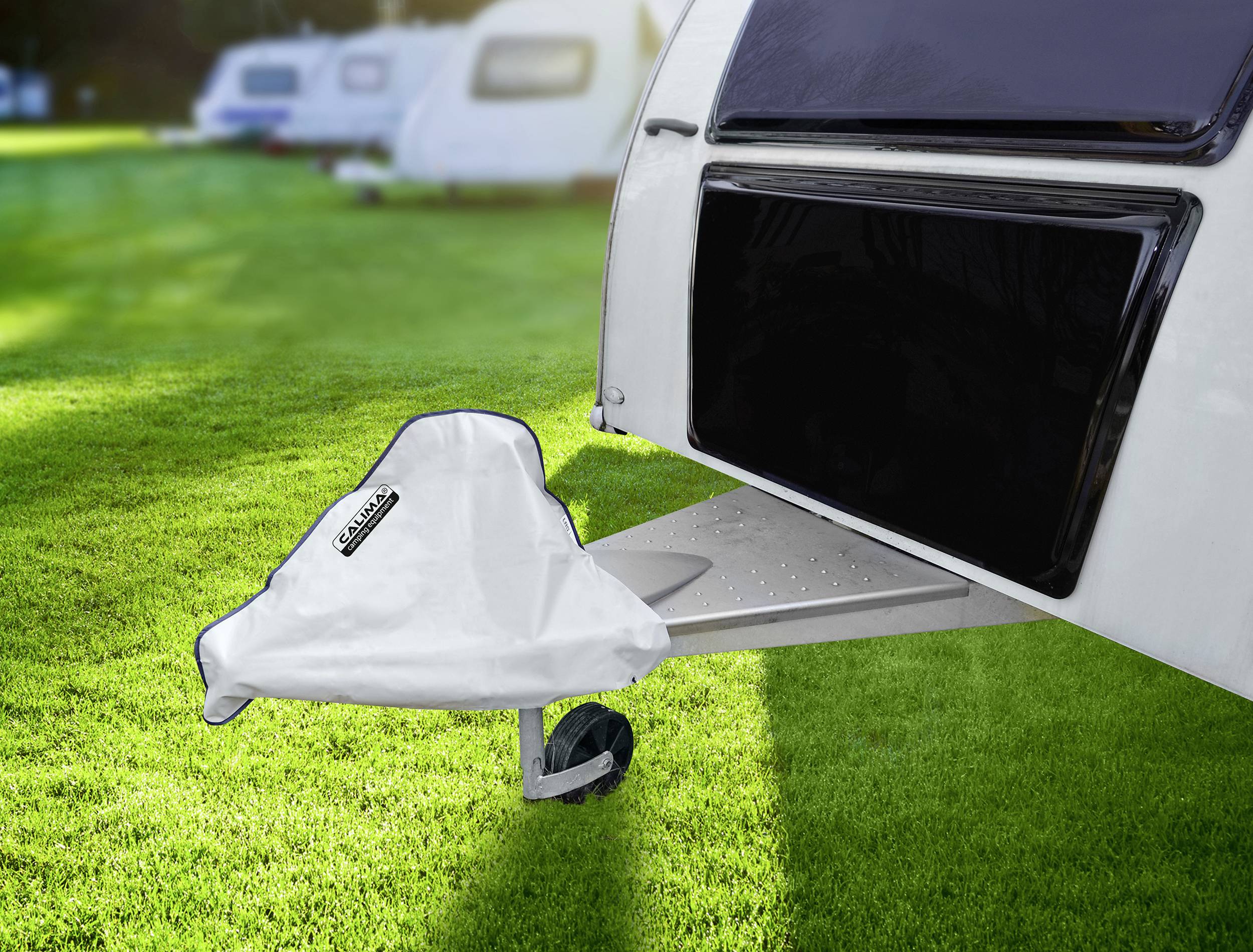 Biały kamper z przykrytą komorą gazową stoi na zielonej trawie. W tle niewyraźnie widać kolejne kampery.