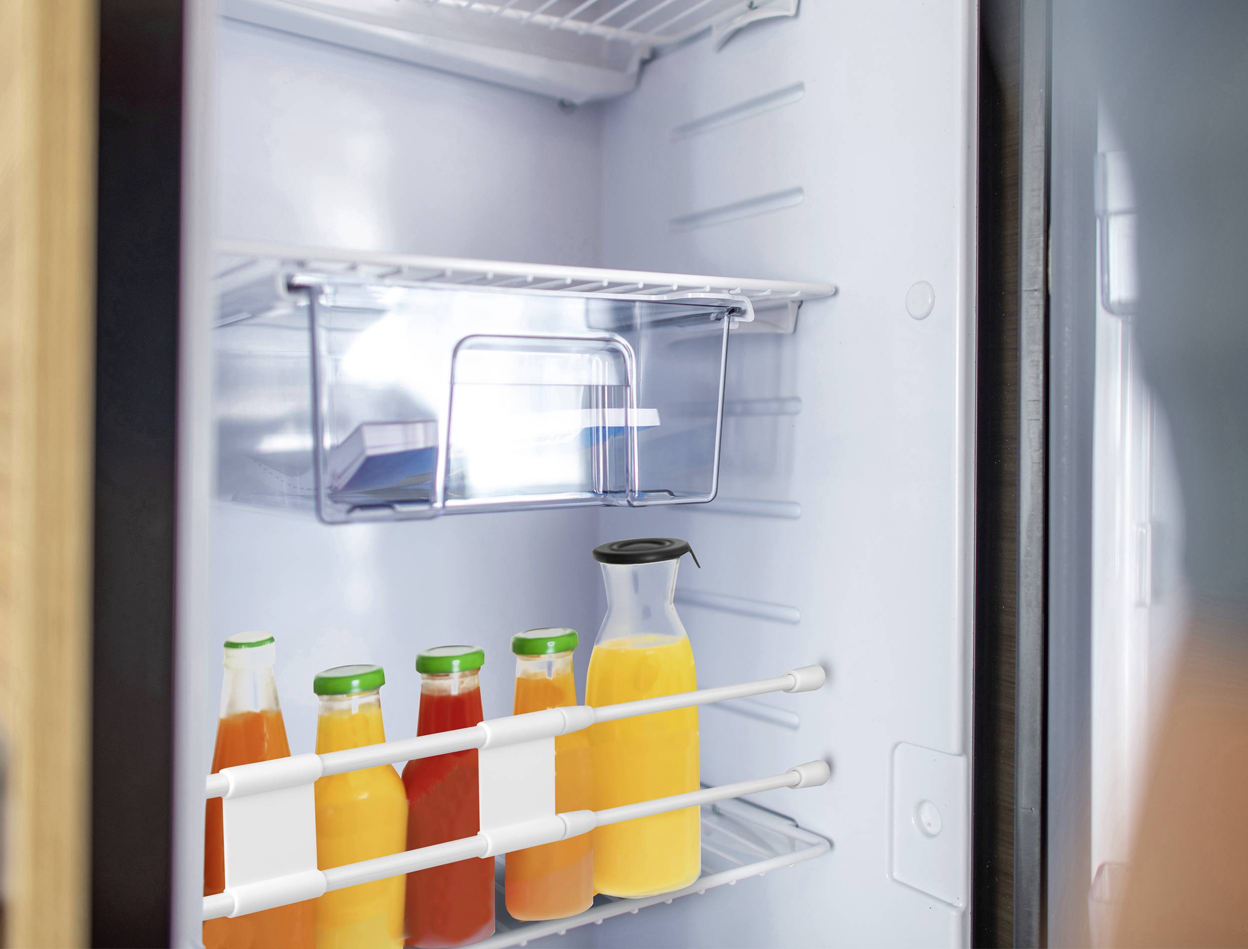 Butelki z sokiem w otwartej lodówce. Cztery mniejsze butelki i jeden większy dzban z sokiem pomarańczowym. Górna półka lodówki pusta.