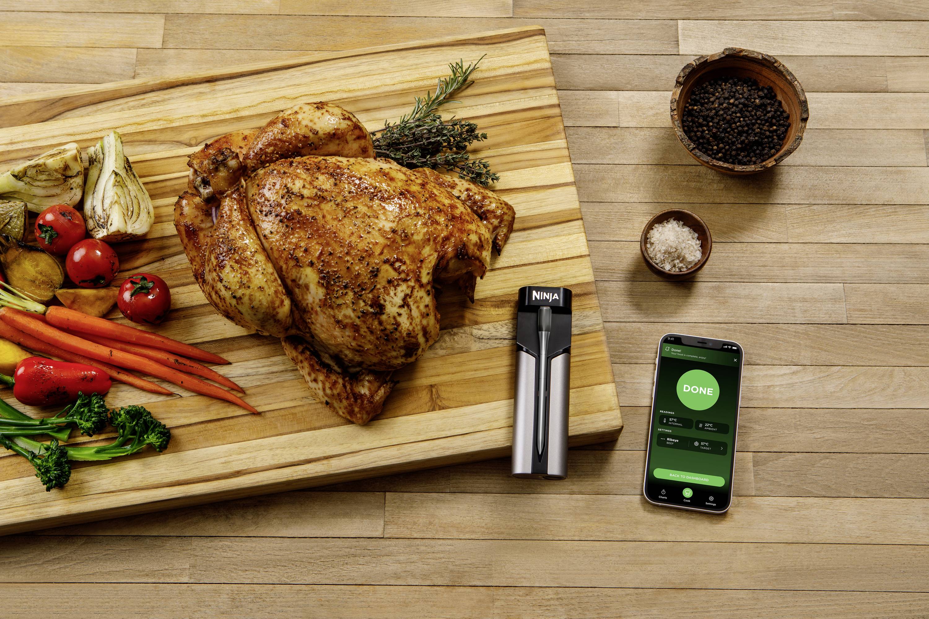 Pieczony kurczak na desce do krojenia z warzywami. Obok leży termometr marki 'Ninja'. Smartfon wyświetla napis 'Gotowe'.