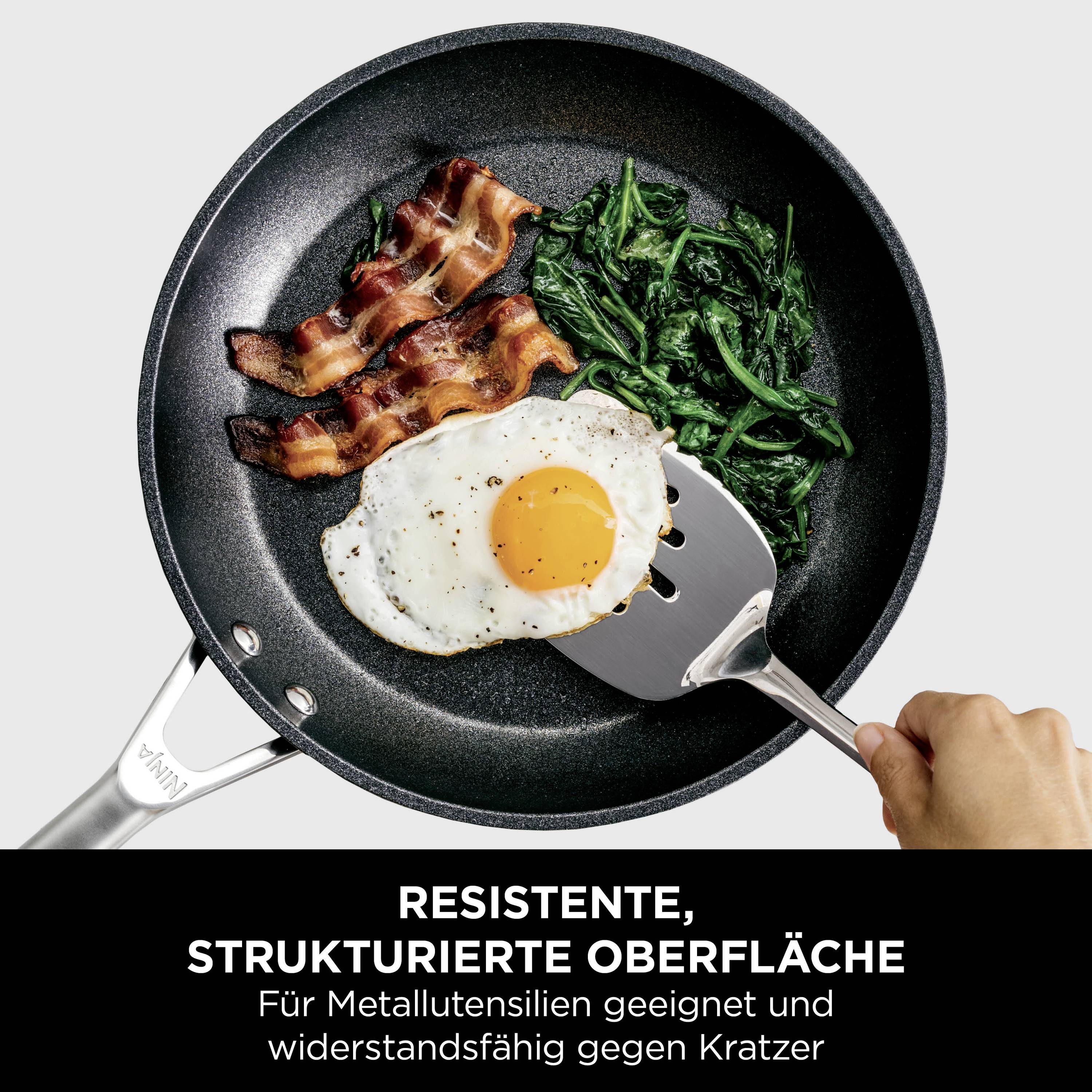 Ręka trzyma patelnię z boczkiem, szpinakiem i jajkiem sadzonym. Tekst: 'Odporna, strukturalna powierzchnia, odpowiednia do przyborów metalowych, odporna na zarysowania'.