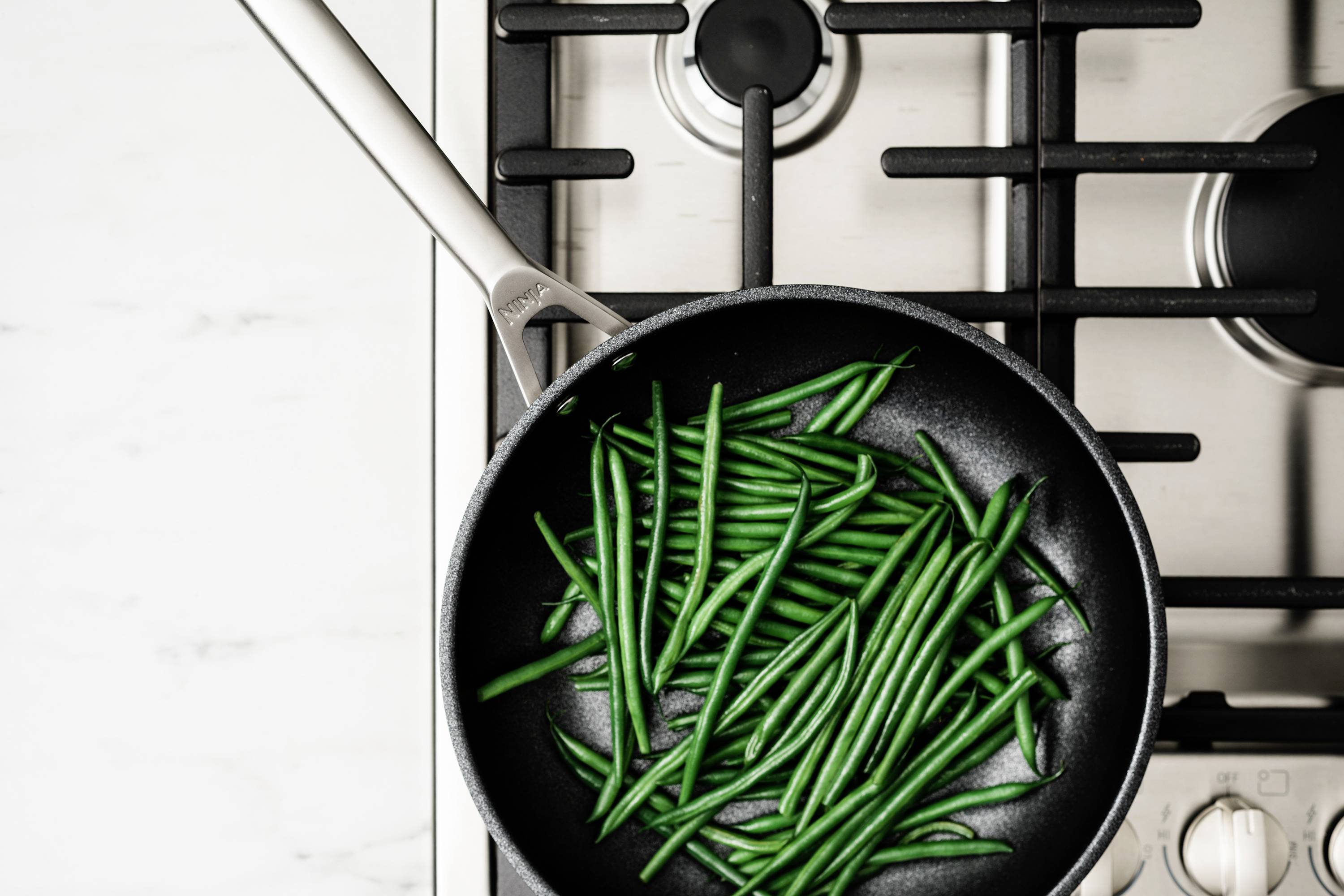 Czarna patelnia z zielonymi fasolkami na kuchence. Fasolki są równomiernie rozłożone i częściowo przyrumienione.
