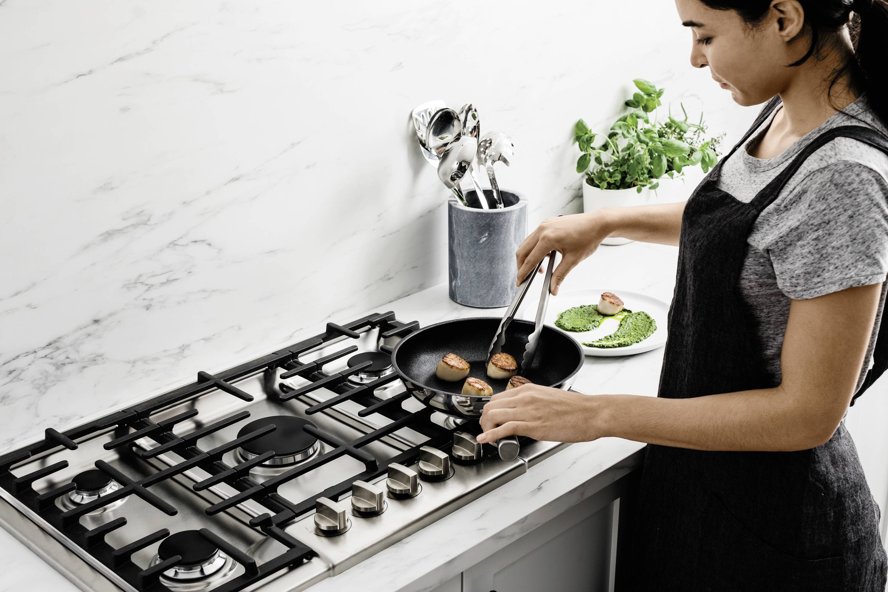 Osoba gotuje przegrzebki na patelni na kuchence, obok znajduje się talerz z brokułami. W tle widoczne są przybory kuchenne i zioła.