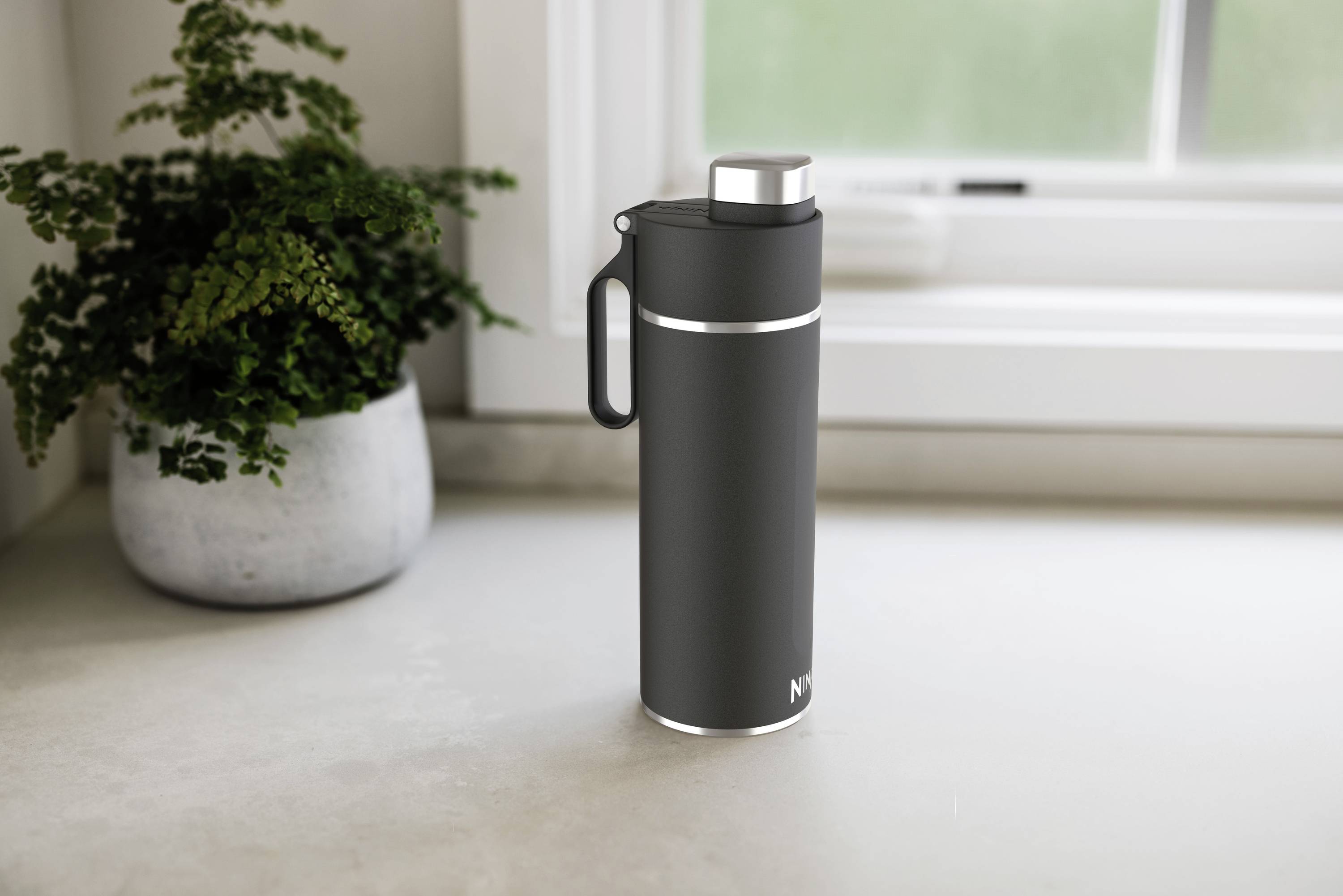 Czarny termos stoi na blacie kuchennym przed oknem, obok doniczki z rośliną.