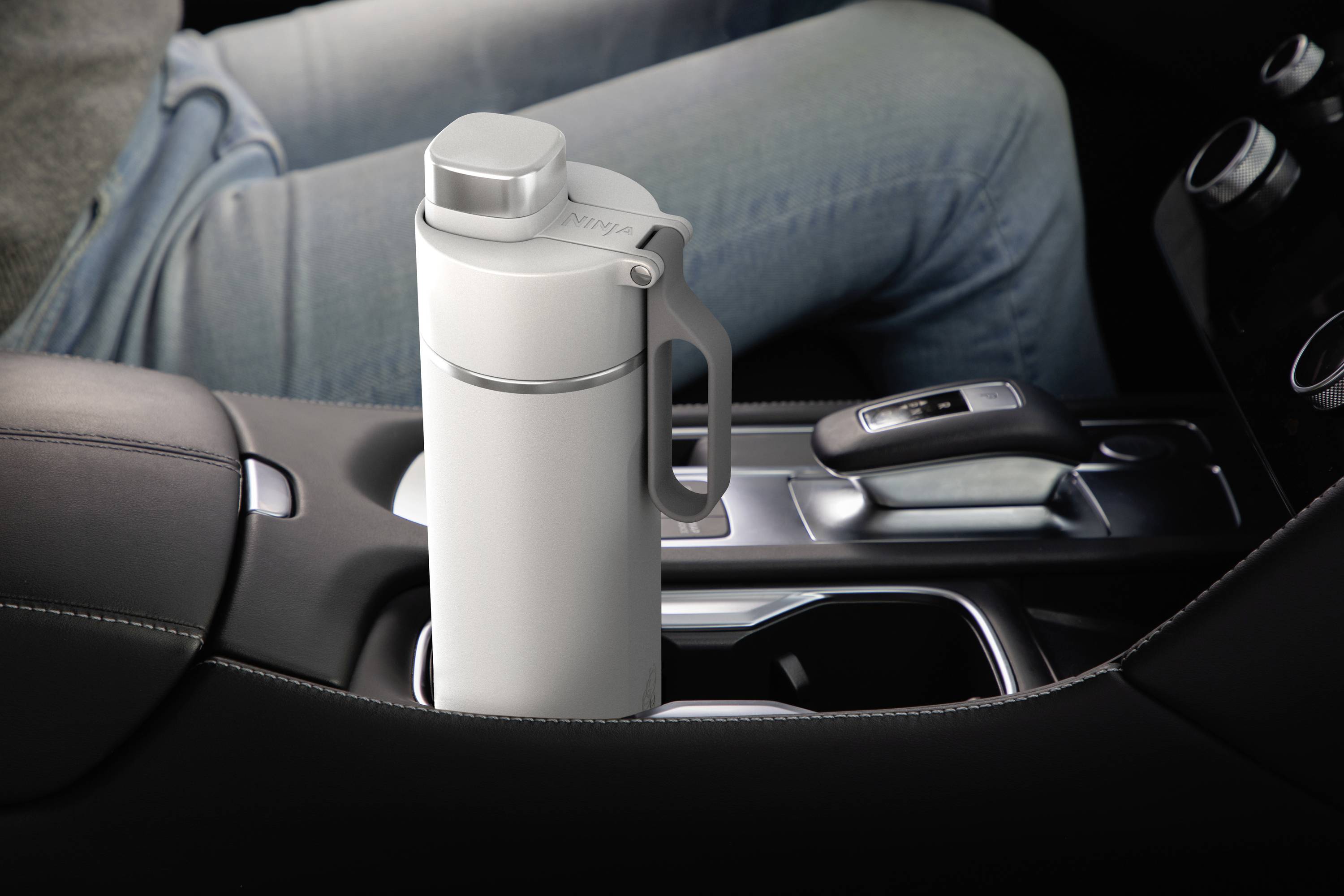 Biała termoska stoi w uchwycie na napoje w samochodzie, obok dźwigni zmiany biegów. Osoba ubrana jest w dżinsy.