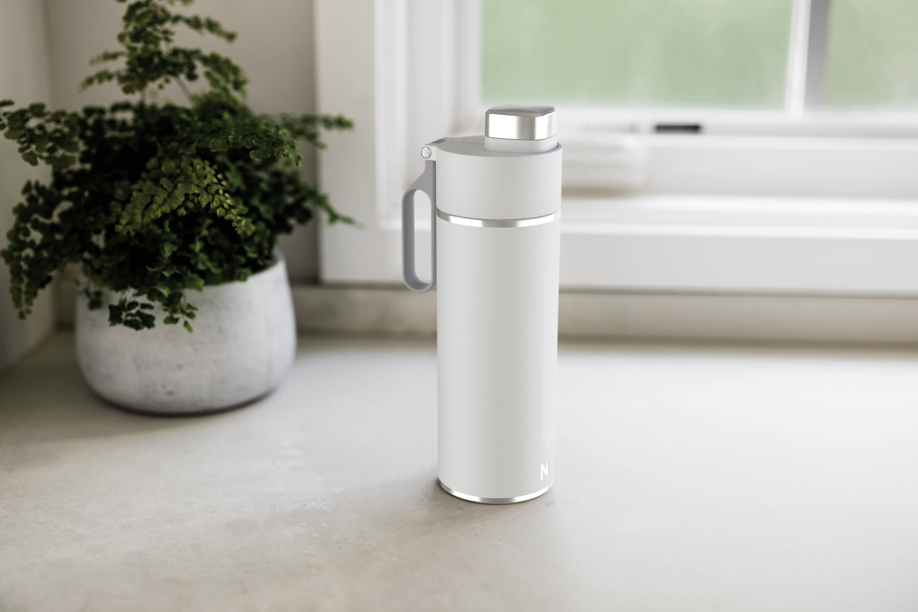 Biała termobutelka stoi na blacie kuchennym przed oknem, obok małej rośliny doniczkowej.