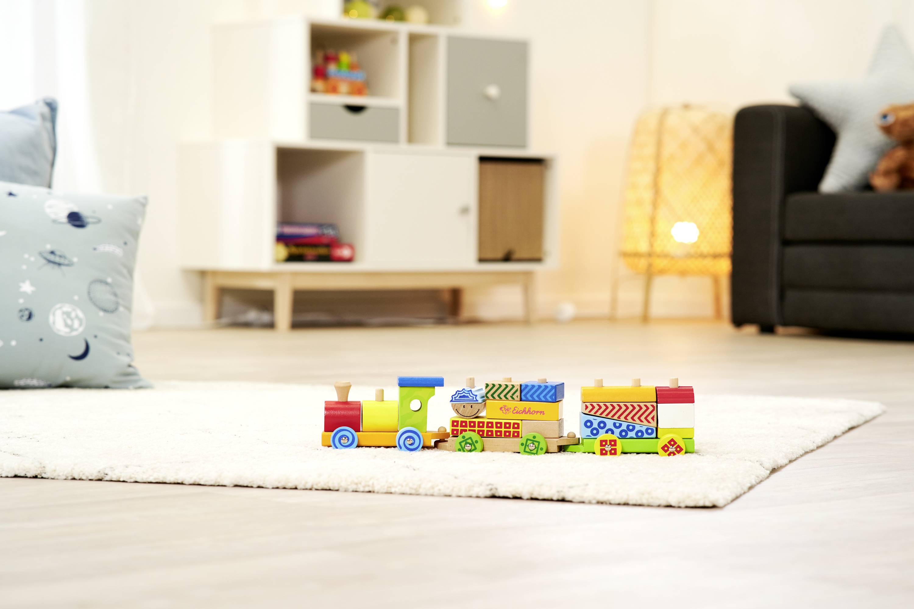 Drewniany pociąg zabawkowy na białym dywanie w salonie, otoczony nowoczesnym, jasnym wystrojem i kanapą w tle.