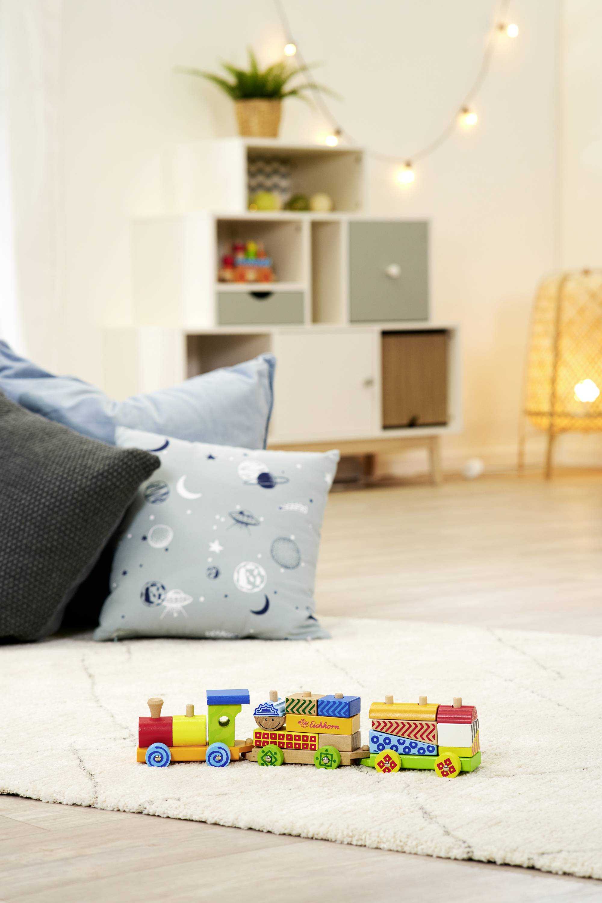 Drewniany pociąg zabawkowy na jasnym dywanie w przytulnym salonie, z poduszkami, regałem i roślinami w tle.