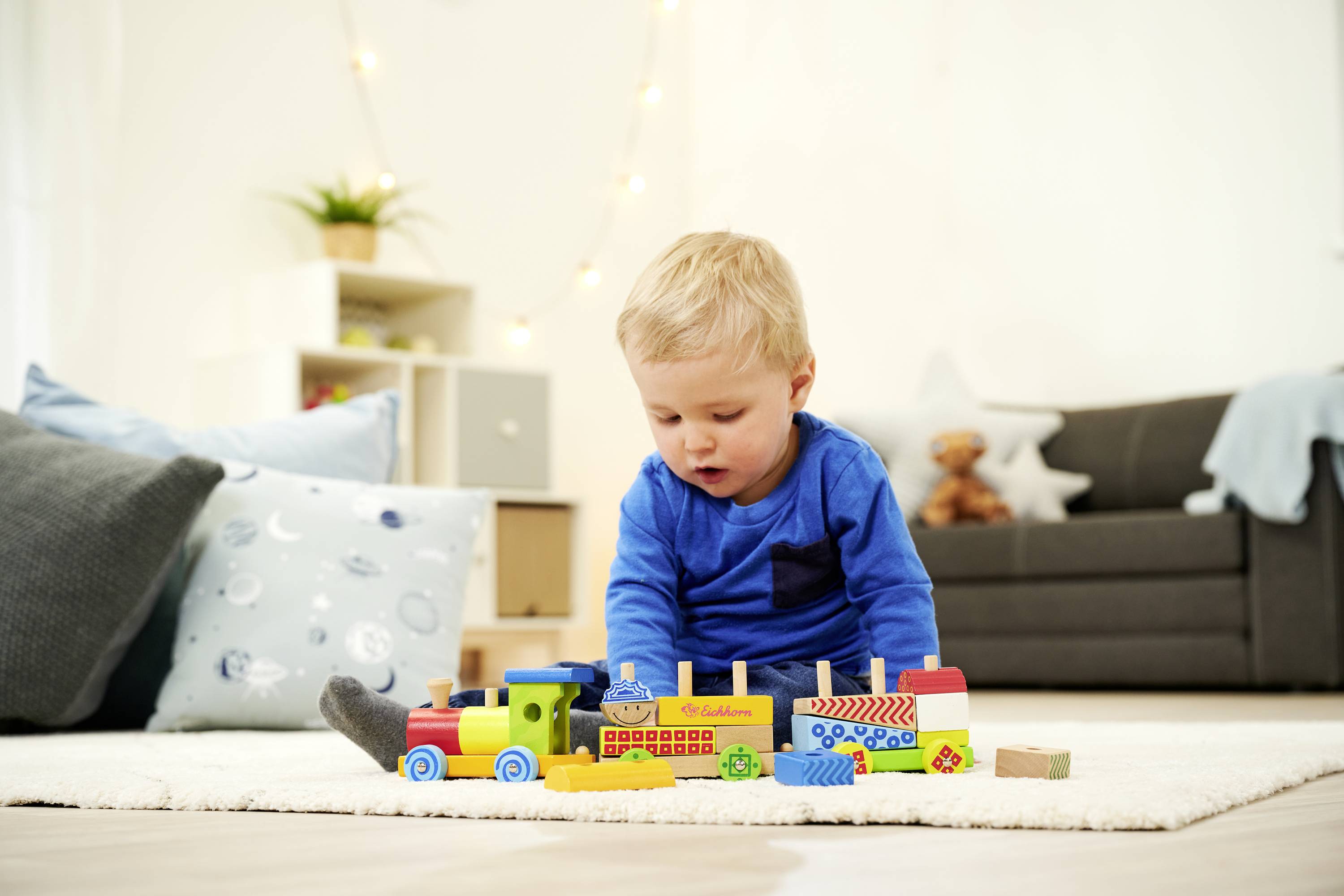 Małe dziecko bawi się kolorową drewnianą kolejką na dywanie w przytulnym salonie. W tle widać sofę i pudła.