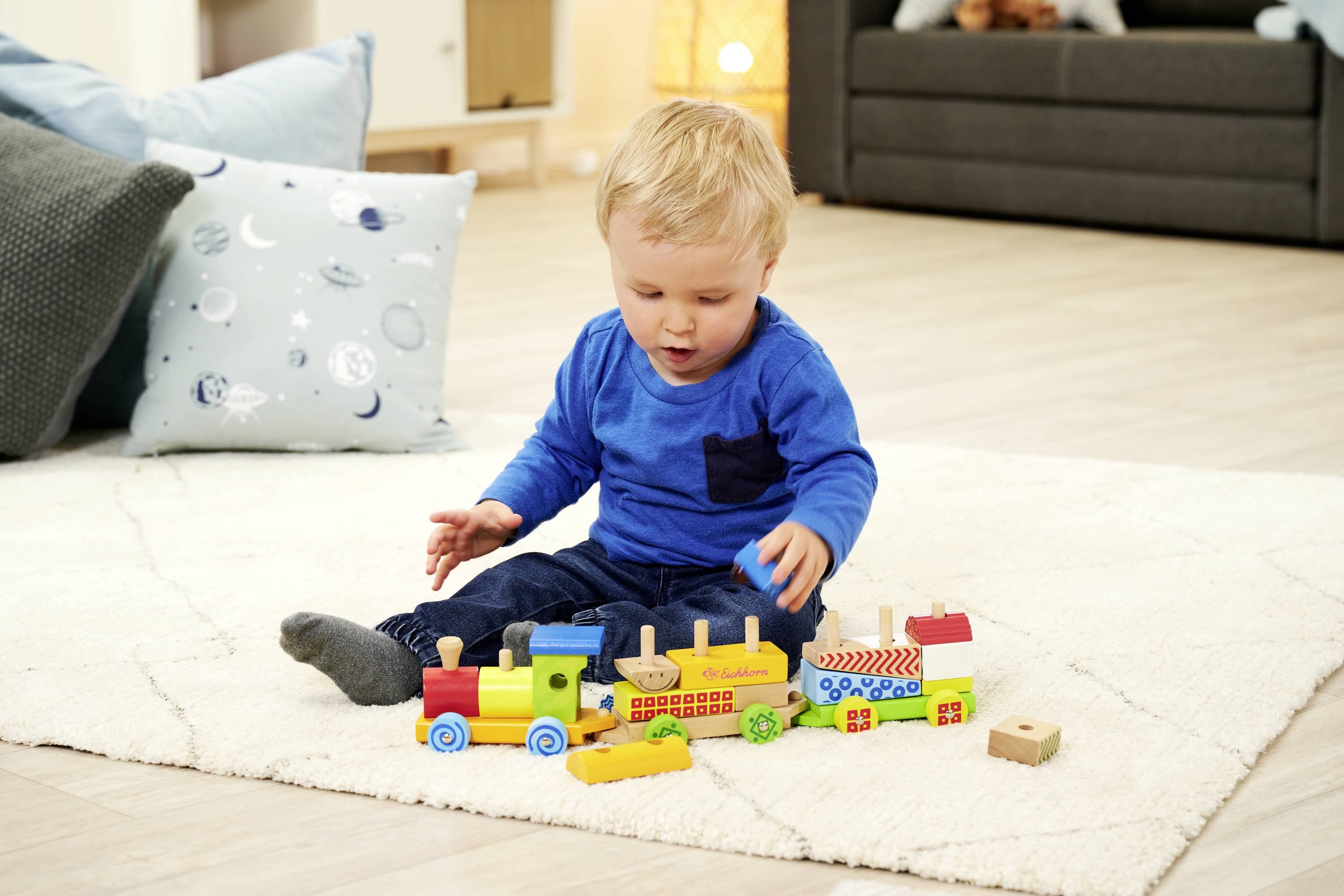 Małe dziecko siedzi na dywanie i bawi się kolorową drewnianą kolejką. W tle widać salon z kanapą i poduszkami.