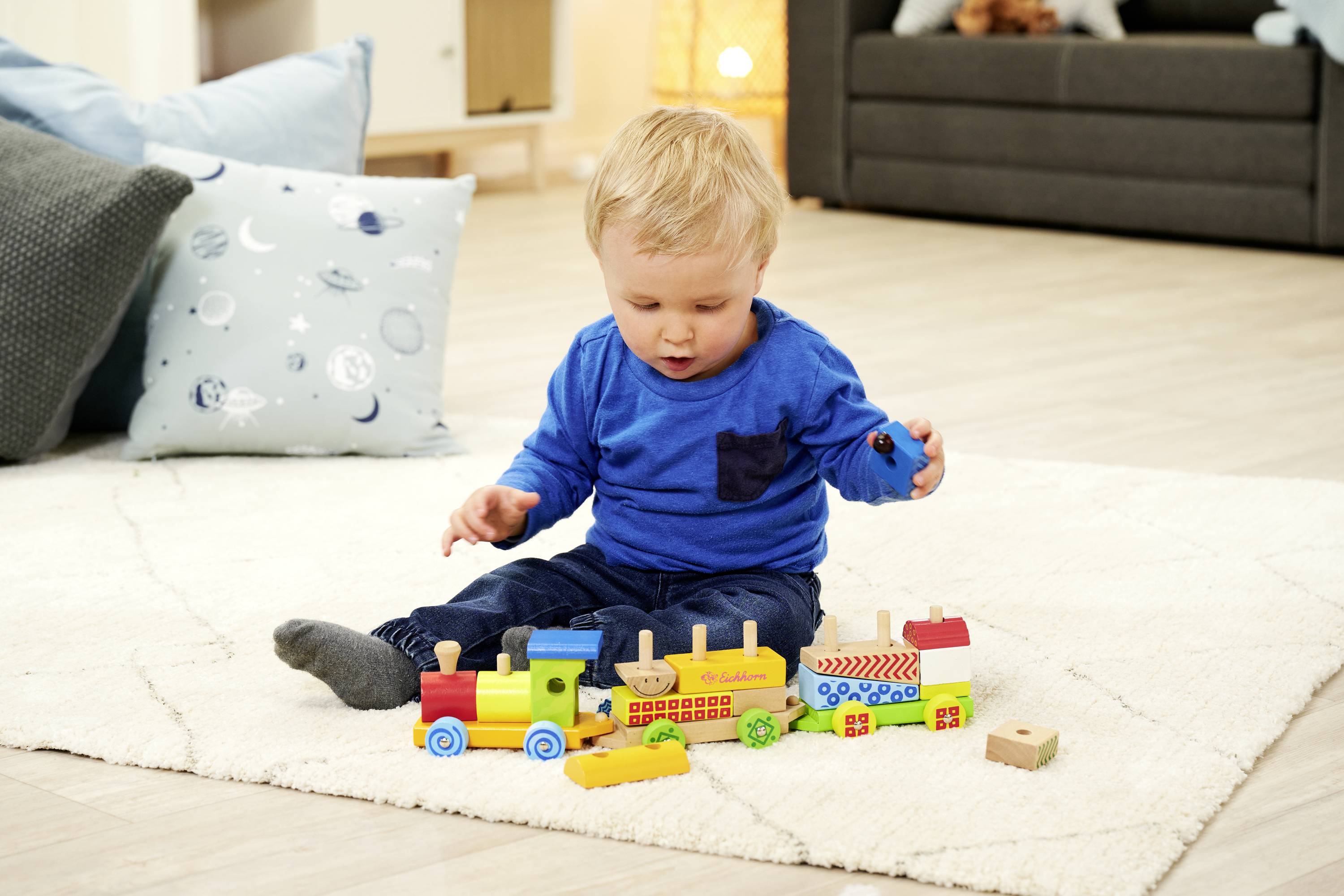 Małe dziecko bawi się kolorową drewnianą kolejką na kremowym dywanie, otoczone poduszkami w przytulnym salonie.