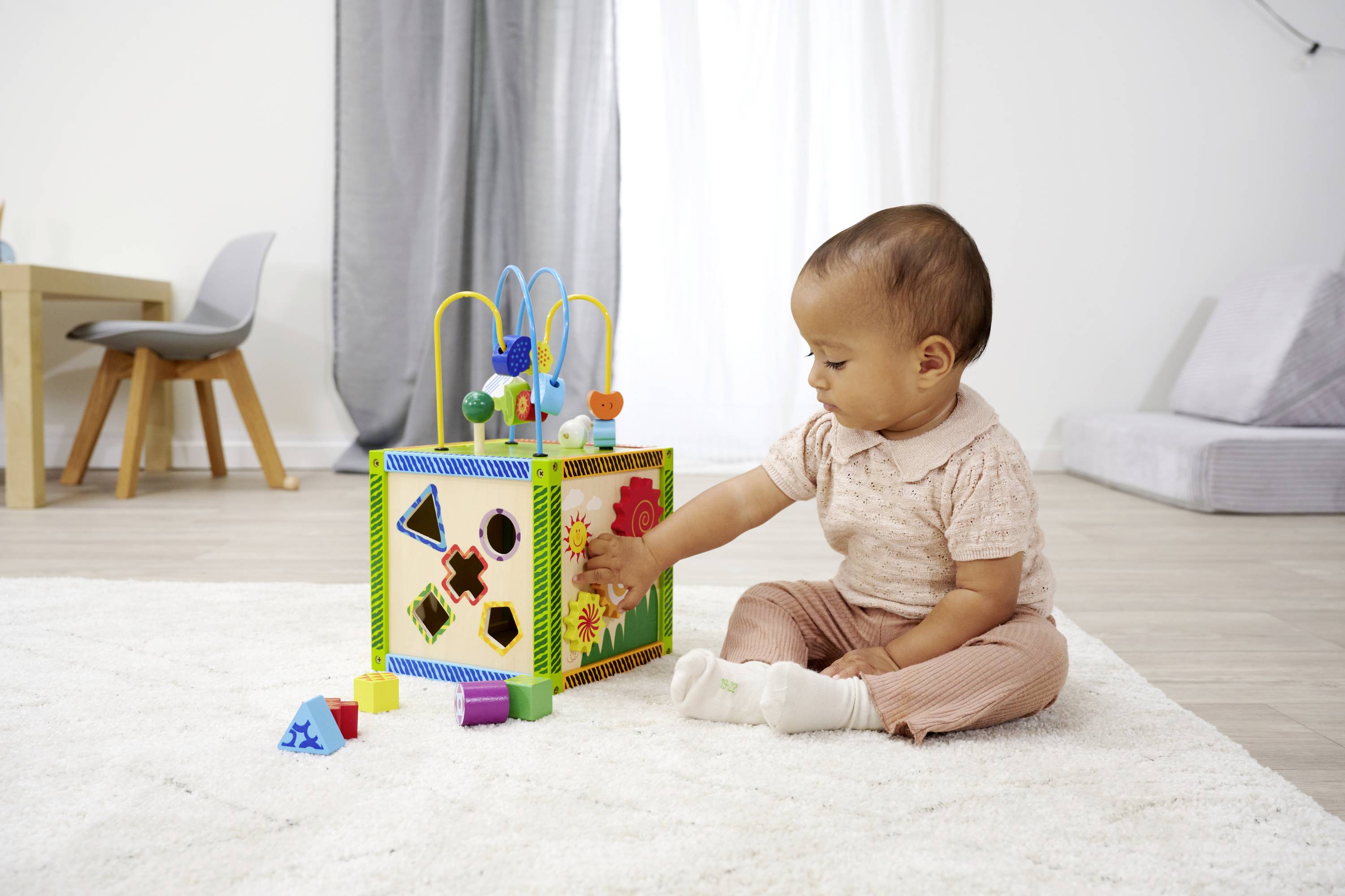 Małe dziecko bawi się na dywanie kolorową kostką edukacyjną z kształtami i zębatkami w jasnym pomieszczeniu.