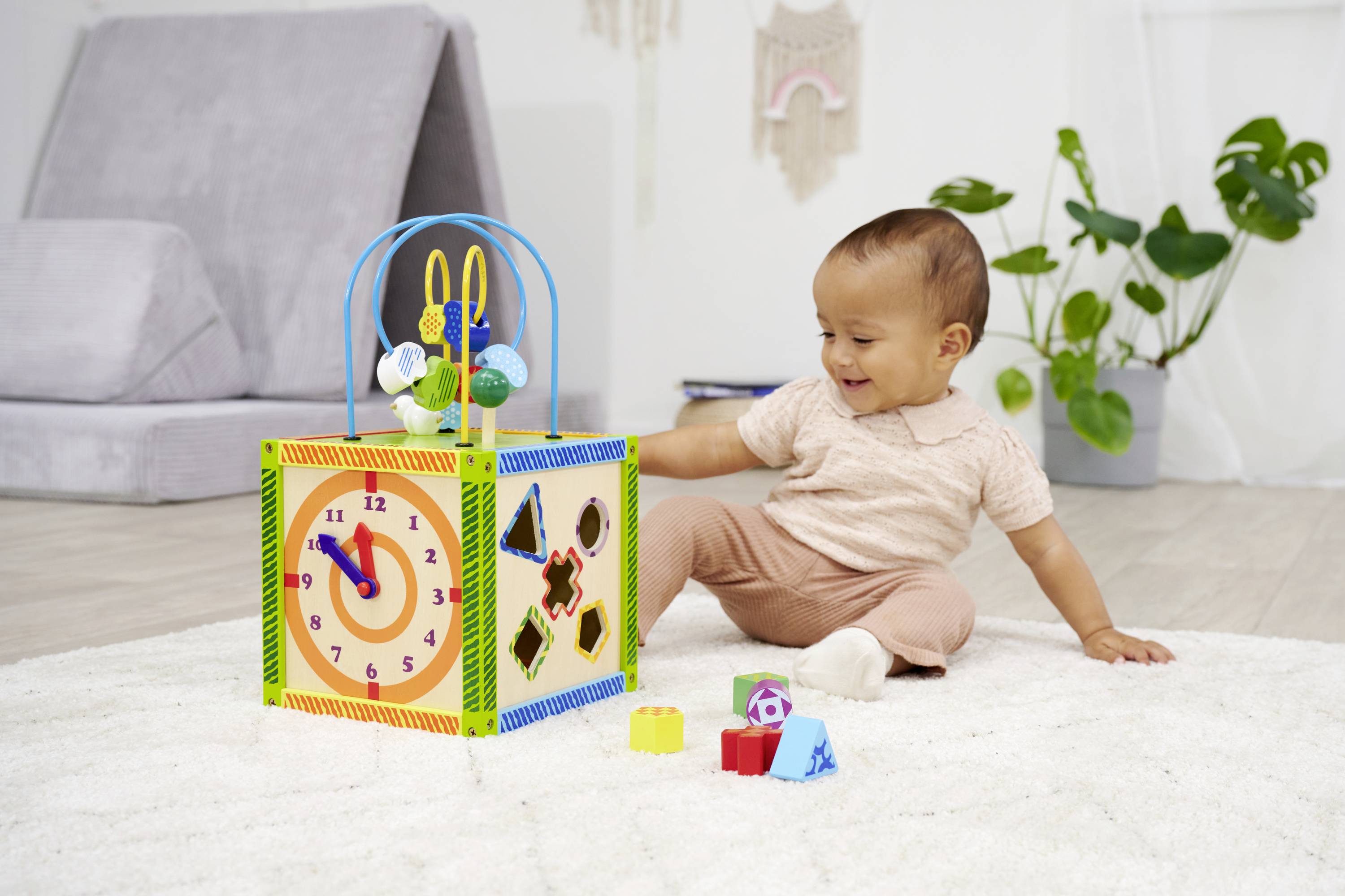 Uśmiechnięte dziecko siedzi na dywanie i bawi się wielokolorową kostką edukacyjną z różnymi kształtami, kolorami i zegarem.