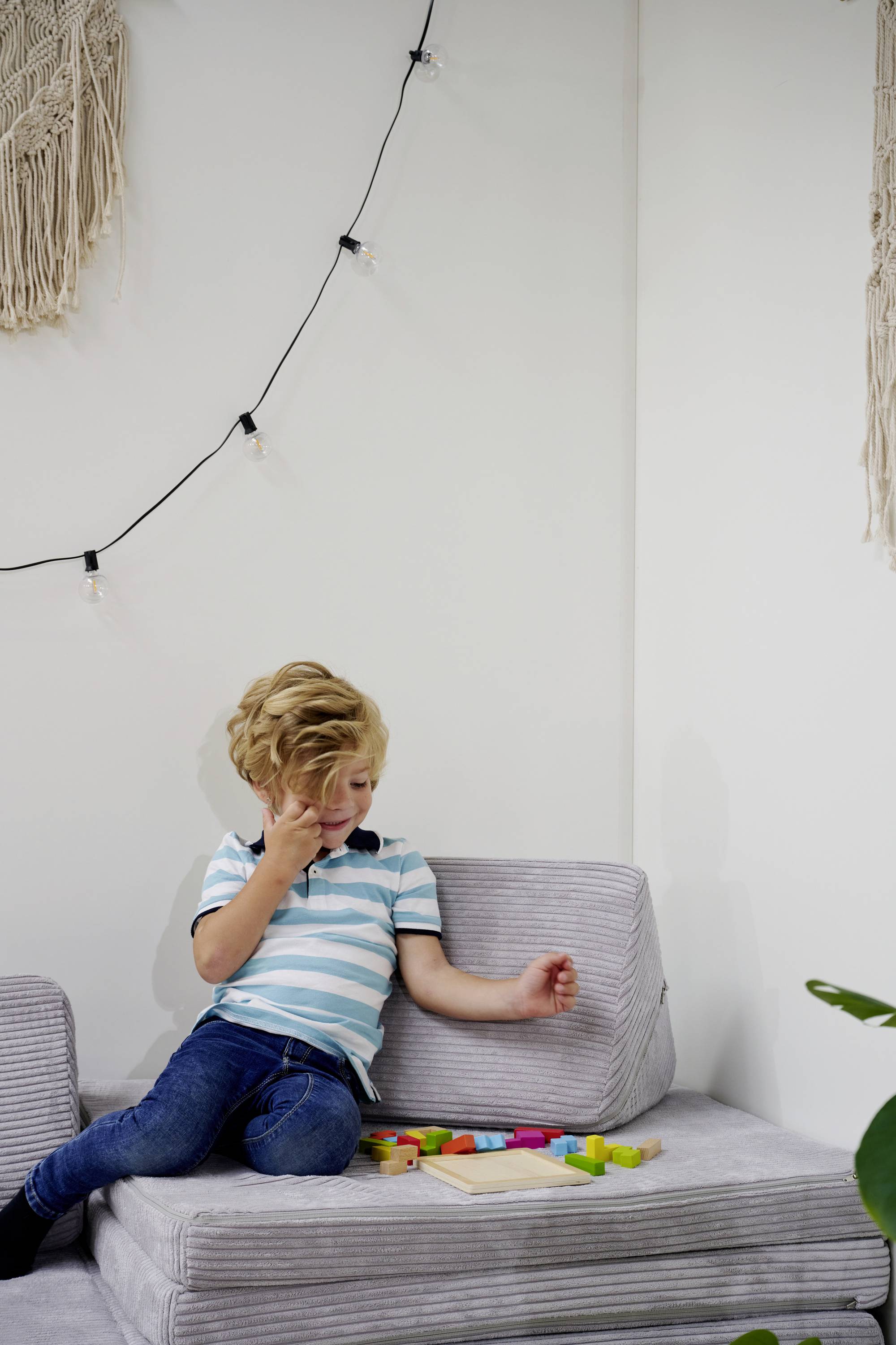 Dziecko siedzi na szarym fotelu i bawi się kolorowymi drewnianymi klockami. W tle wiszą girlandy świetlne i dekoracje ścienne.