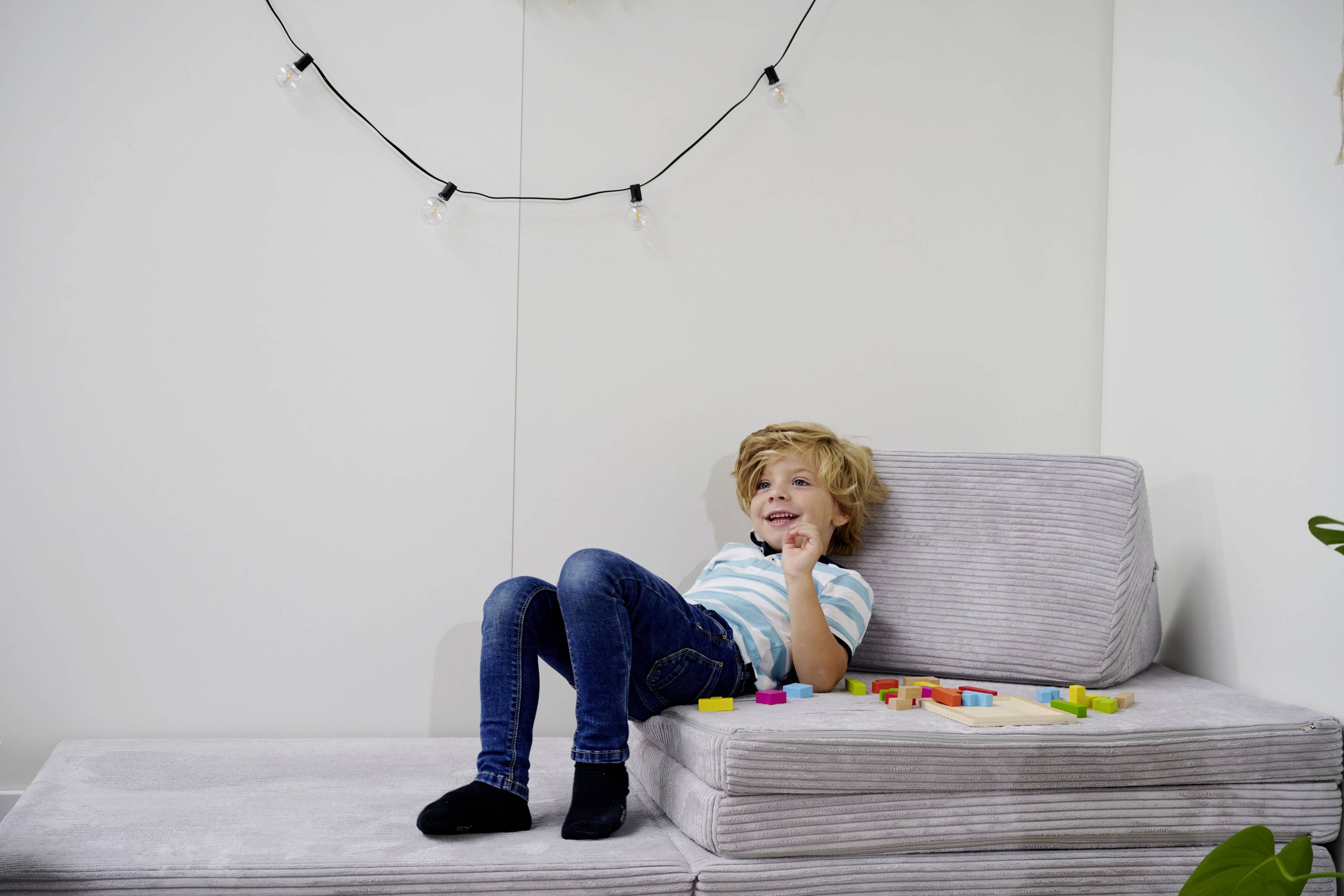 Radosne dziecko siedzi na szarej sofie, otoczone kolorowymi klockami. W tle wisi girlanda świetlna.