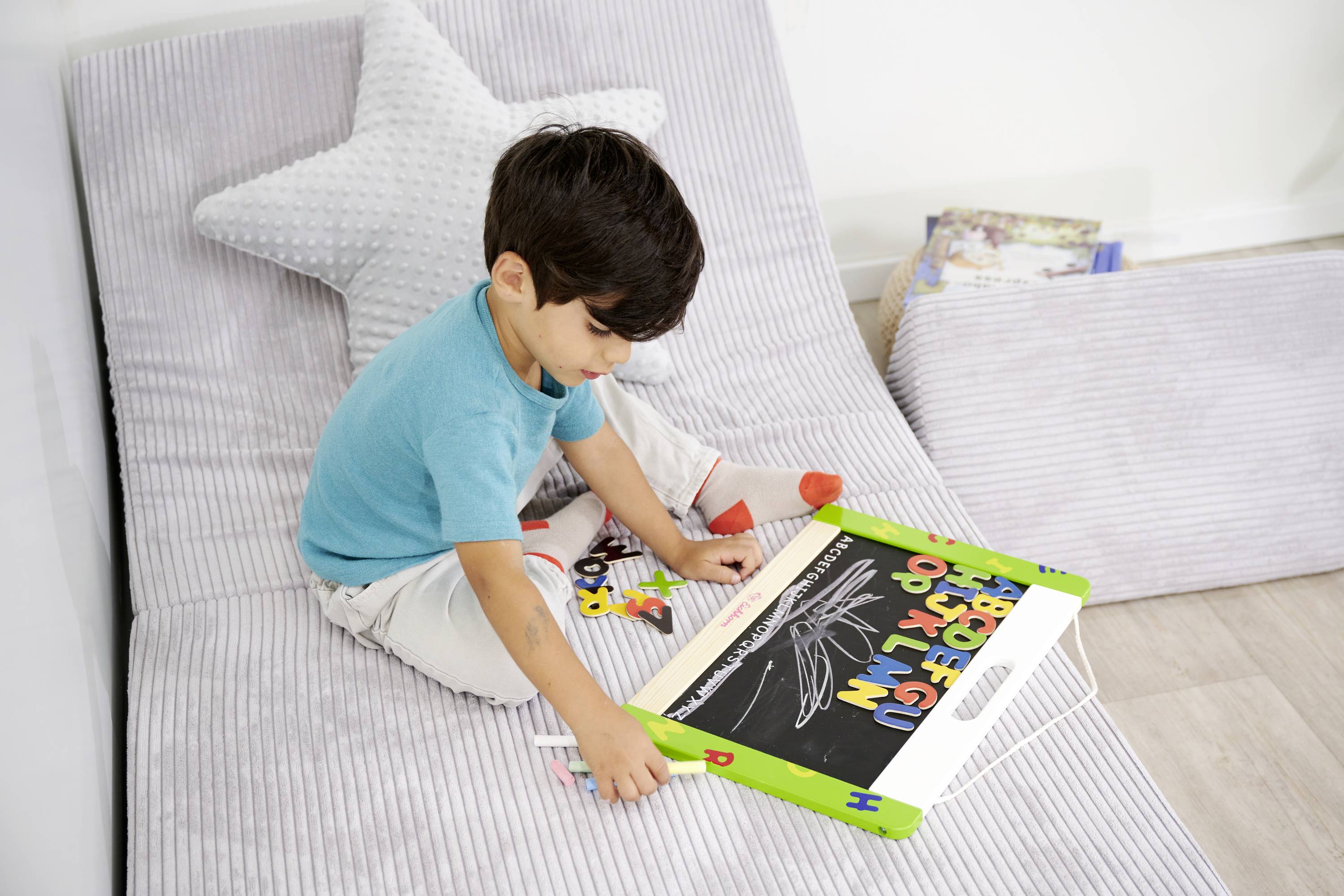 Dziecko siedzi na kanapie i rysuje kredą na małej tablicy, otoczone poduszką w kształcie gwiazdy i zabawkami.