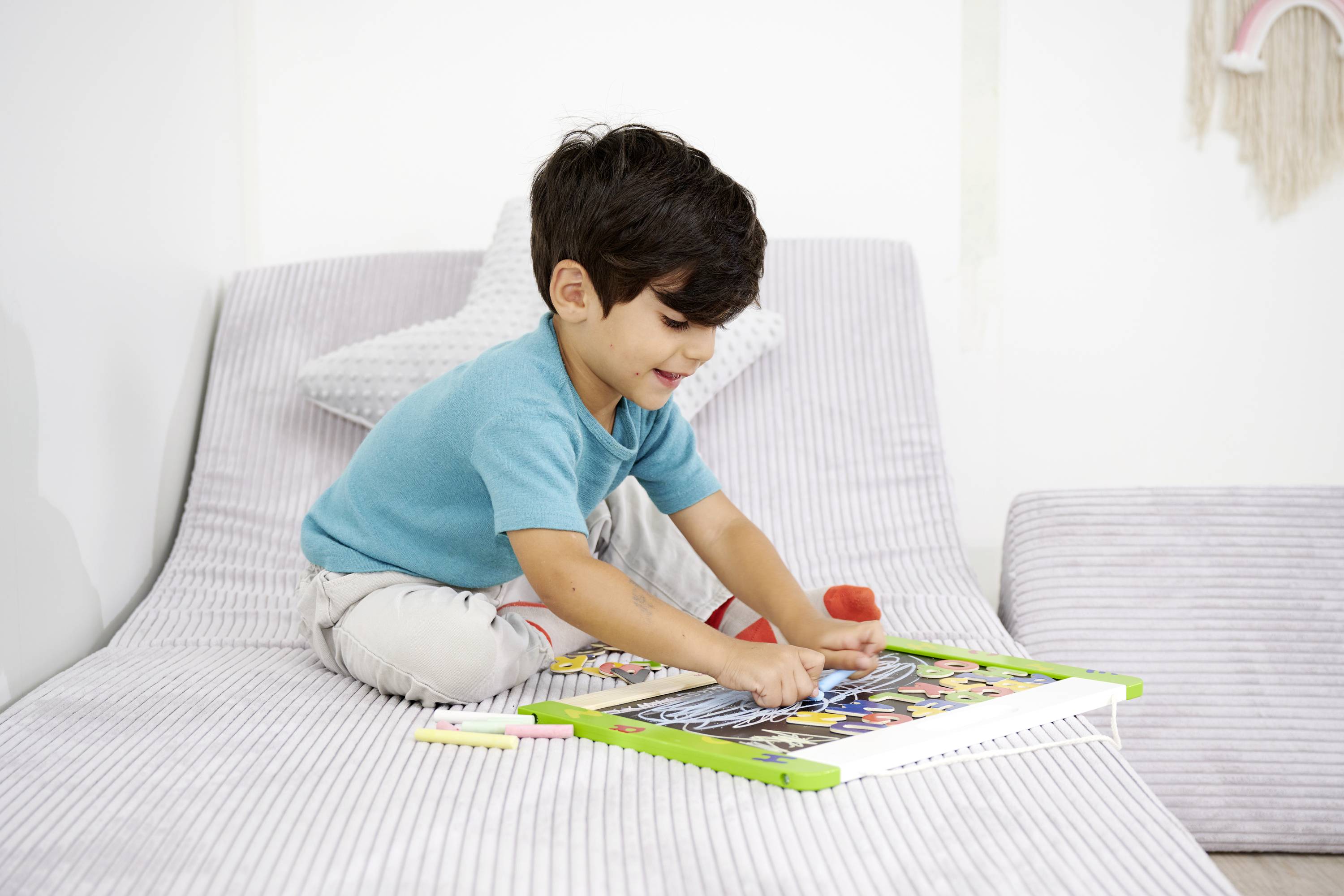 Dziecko siedzi na sofie i rysuje w dużej książce do kolorowania kredkami. Wygląda na bardzo skupione i uśmiechnięte.