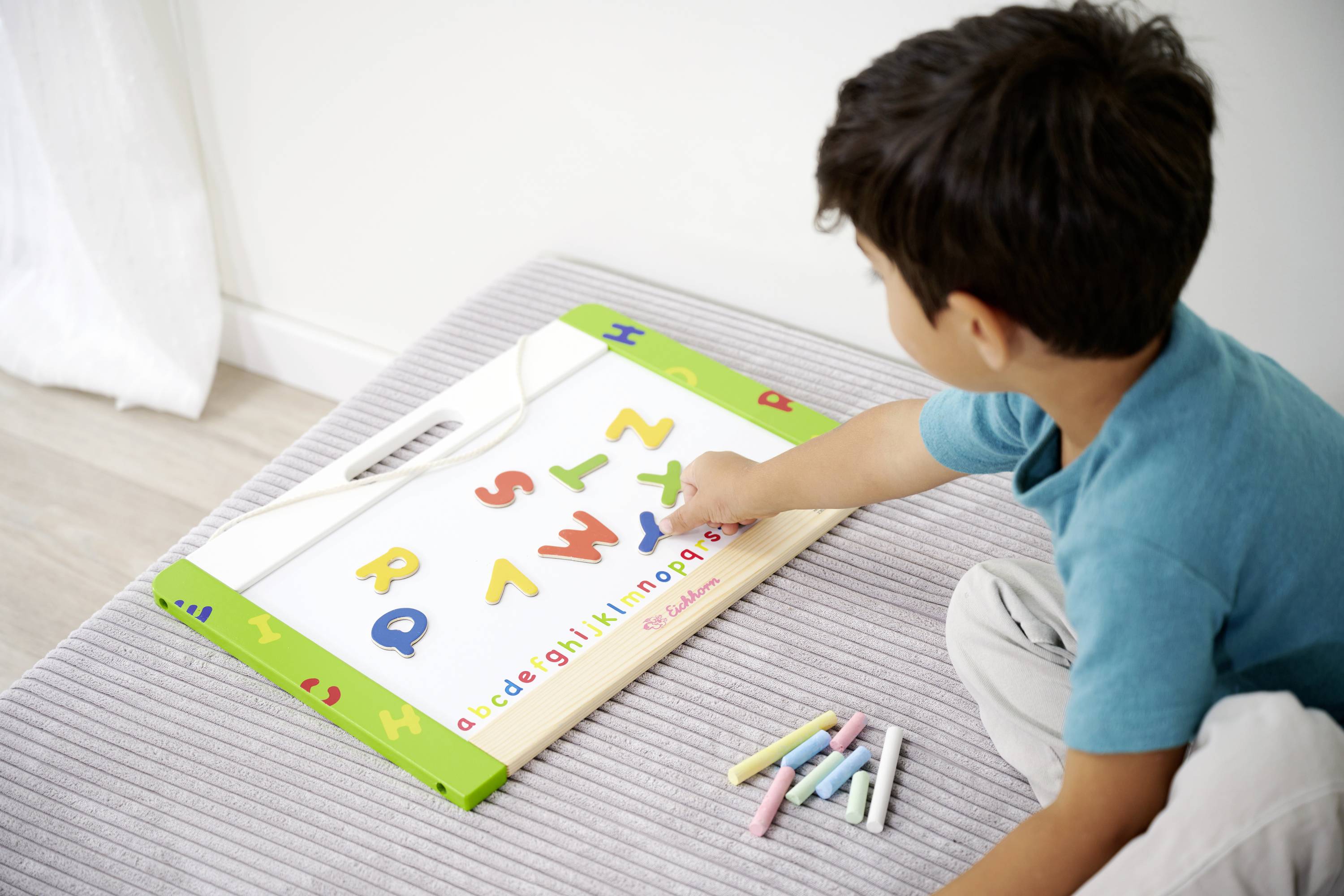Dziecko siedzi na dywanie i bawi się tablicą magnetyczną z kolorowymi literami i cyframi. Obok leży kreda.
