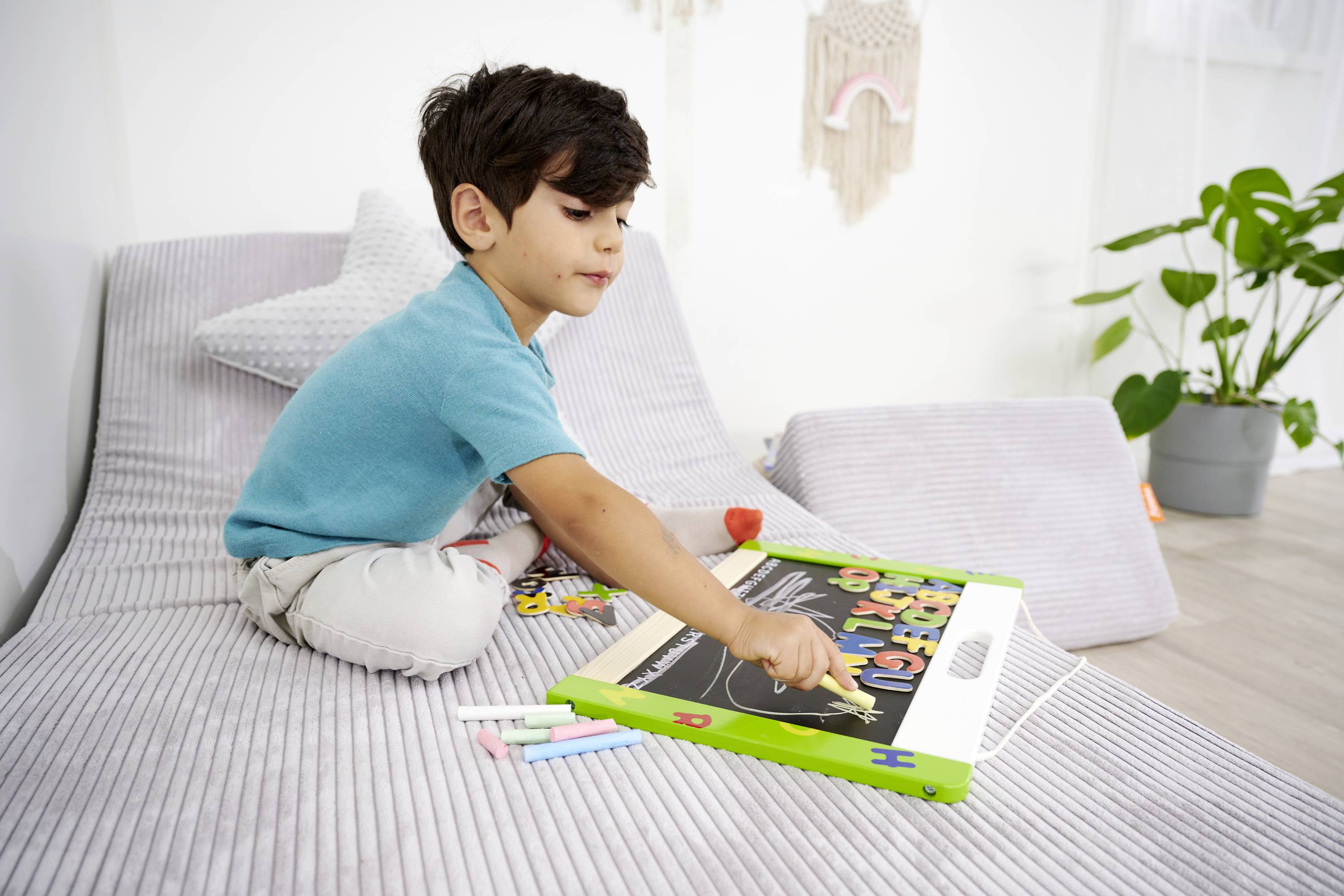 Dziecko siedzi na kanapie i rysuje kredą w małym zeszycie z tablicą. Obok niego leżą kolorowe kawałki kredy. W tle widać roślinę.