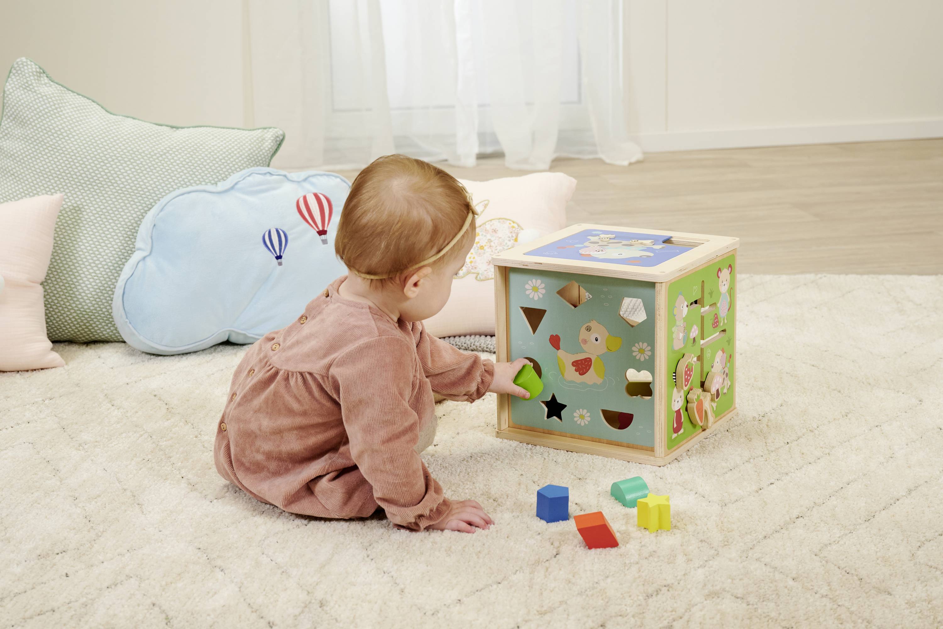 Małe dziecko bawi się na dywanie klockiem interaktywnym, wkładając zielony klocek w odpowiedni otwór. W tle widać poduszki.