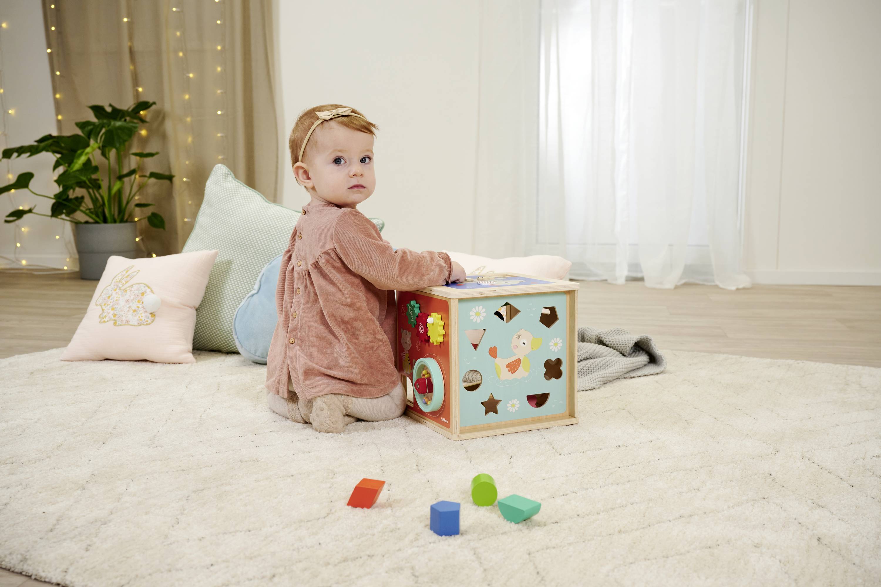 Małe dziecko siedzi na dywanie i bawi się kostką do sortowania kształtów. W tle widać poduszki i roślinę.