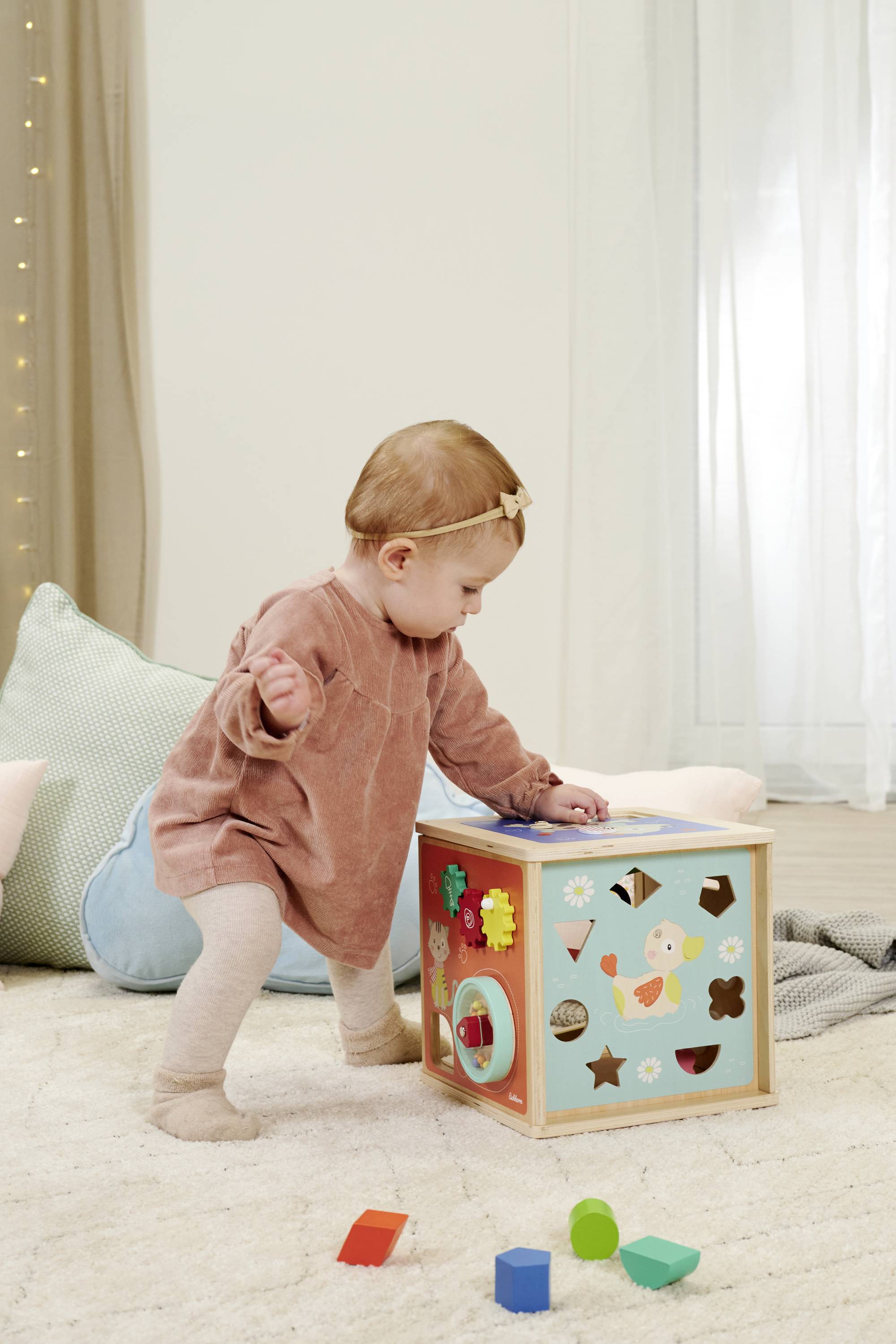 Małe dziecko bawi się kostką zabawkową na dywanie. Kostka ma różne kształty i kolory. W tle widoczne są poduszki.