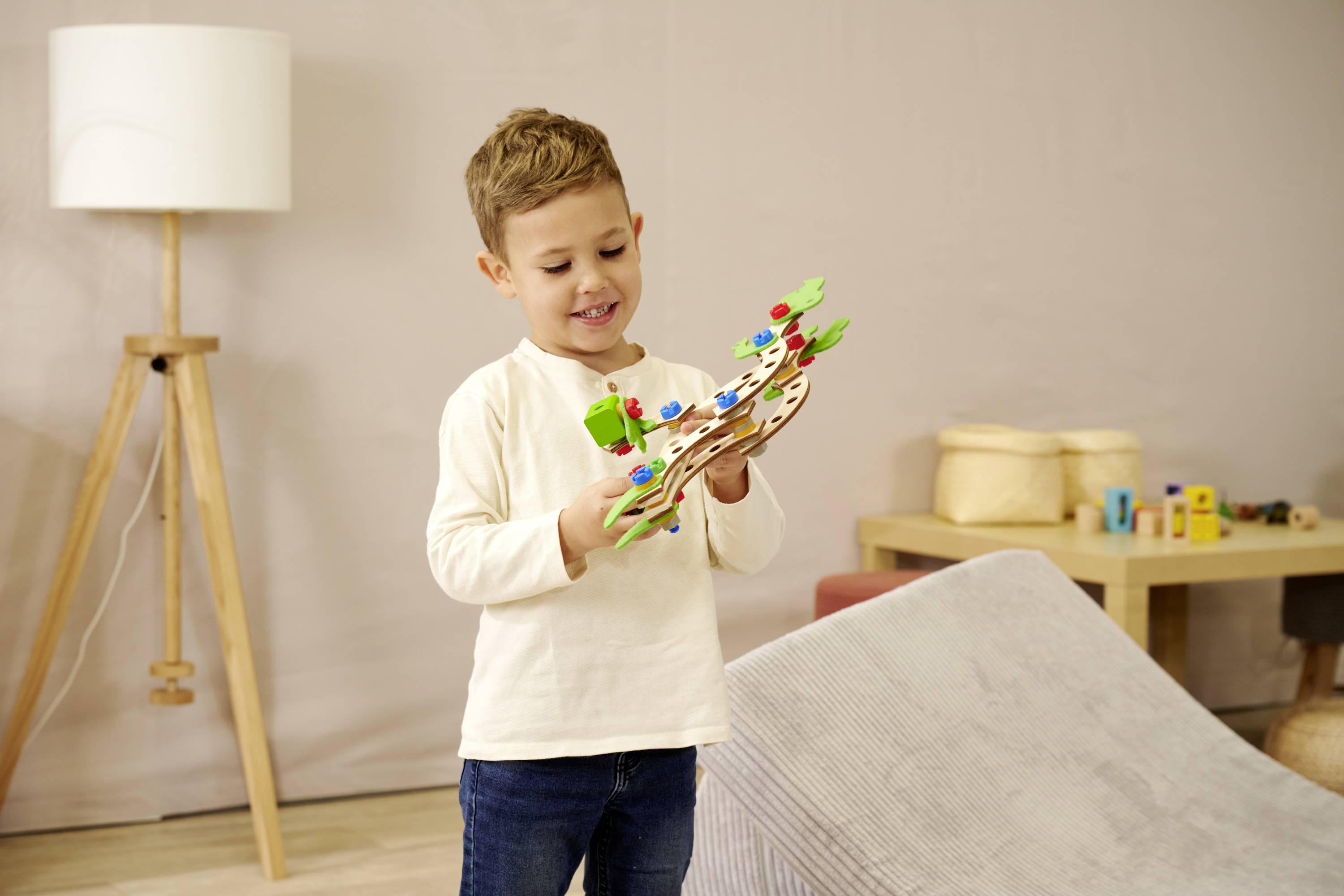 Chłopiec trzyma kolorową drewnianą zabawkę i patrzy na nią. W tle widoczny jest stół z zabawkami, lampa stojąca i sofa.