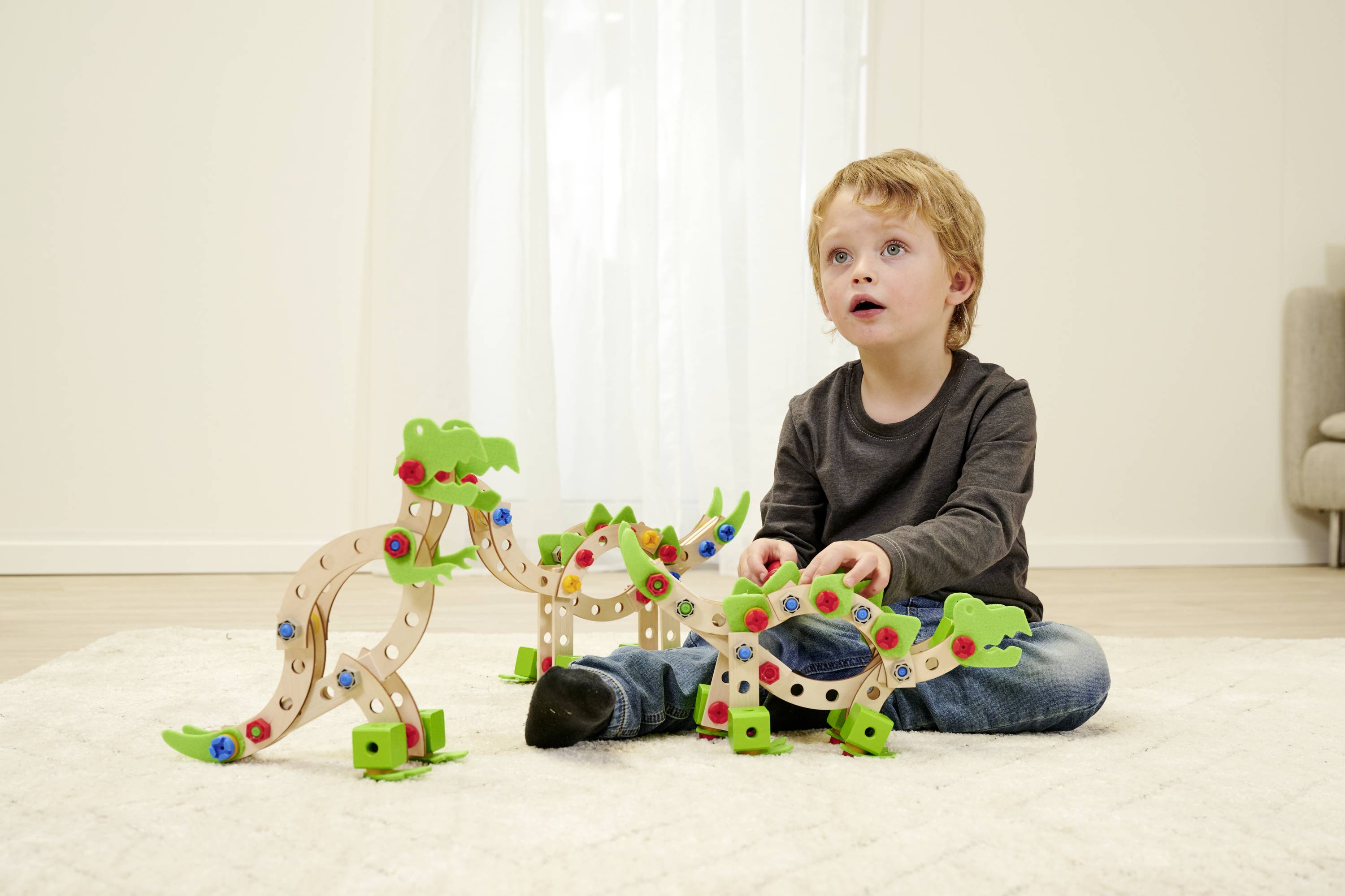 Dziecko siedzi na dywanie i bawi się kolorowymi drewnianymi zabawkami w kształcie dinozaurów w jasnym pomieszczeniu.