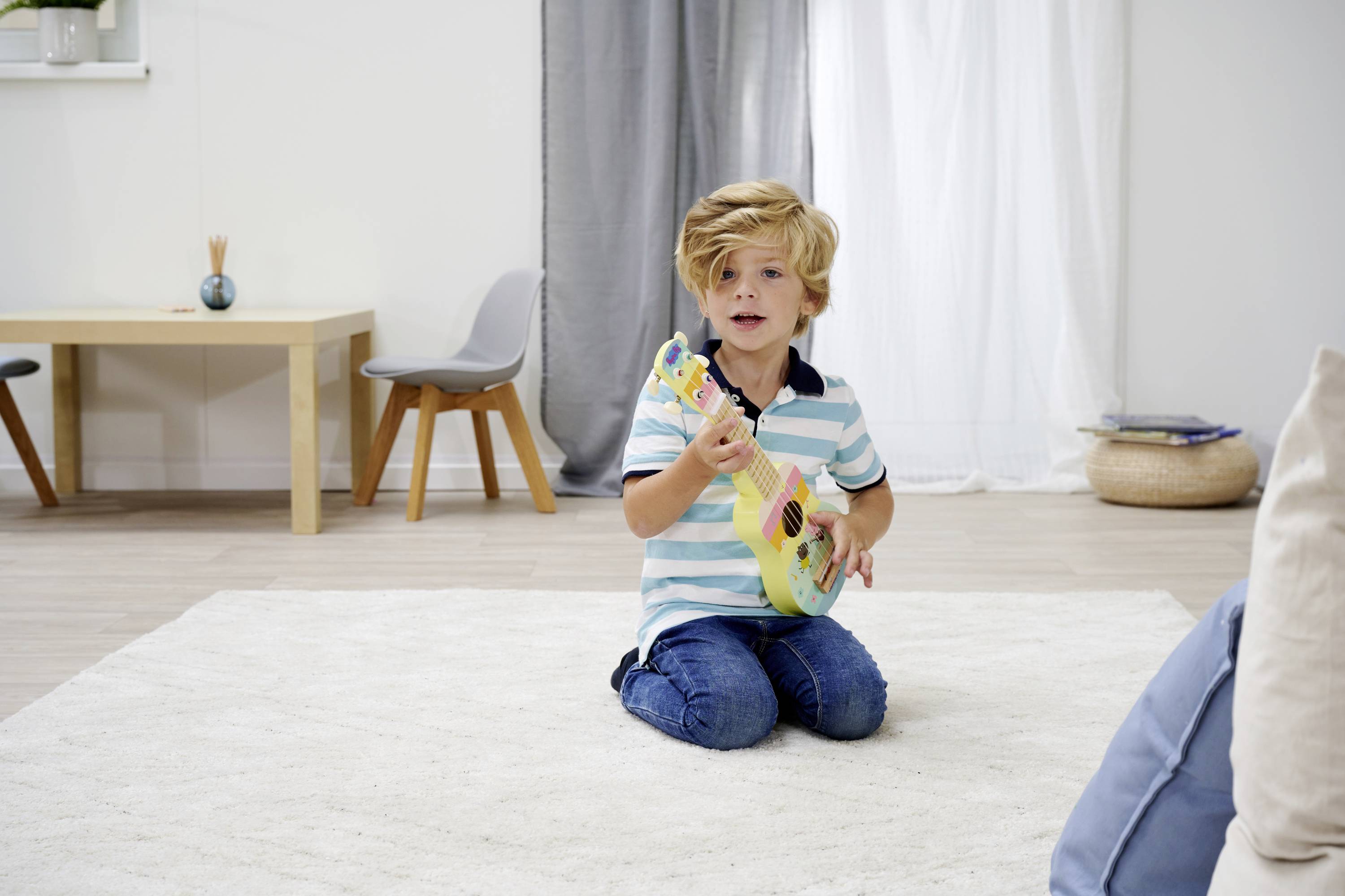 Dziecko siedzi na dywanie i gra na kolorowej ukulele w jasnym salonie, w tle widać stół i krzesła.