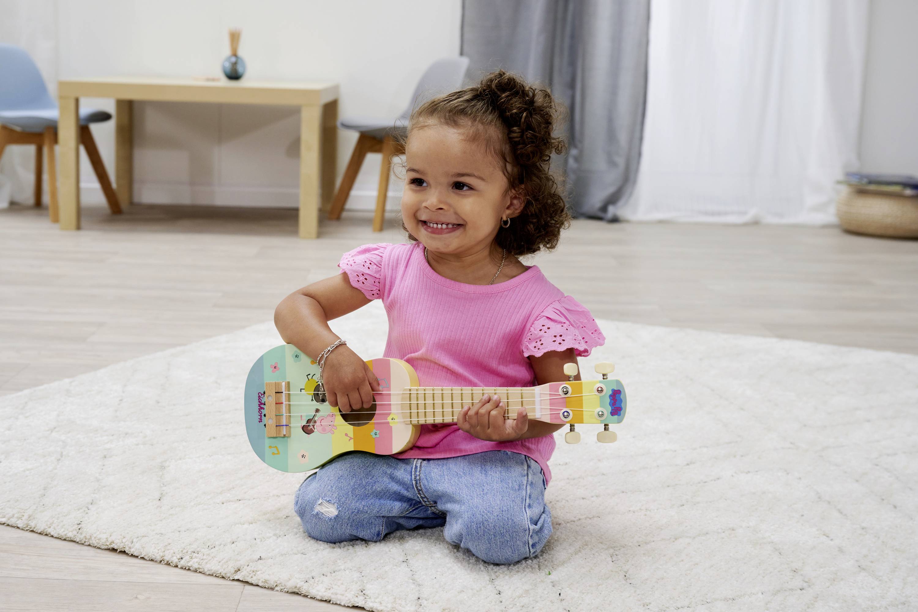Mała dziewczynka w różowym t-shircie i dżinsach siedzi na dywanie i bawi się kolorową, zabawkową ukulele. Uśmiecha się. W tle stoi stół.